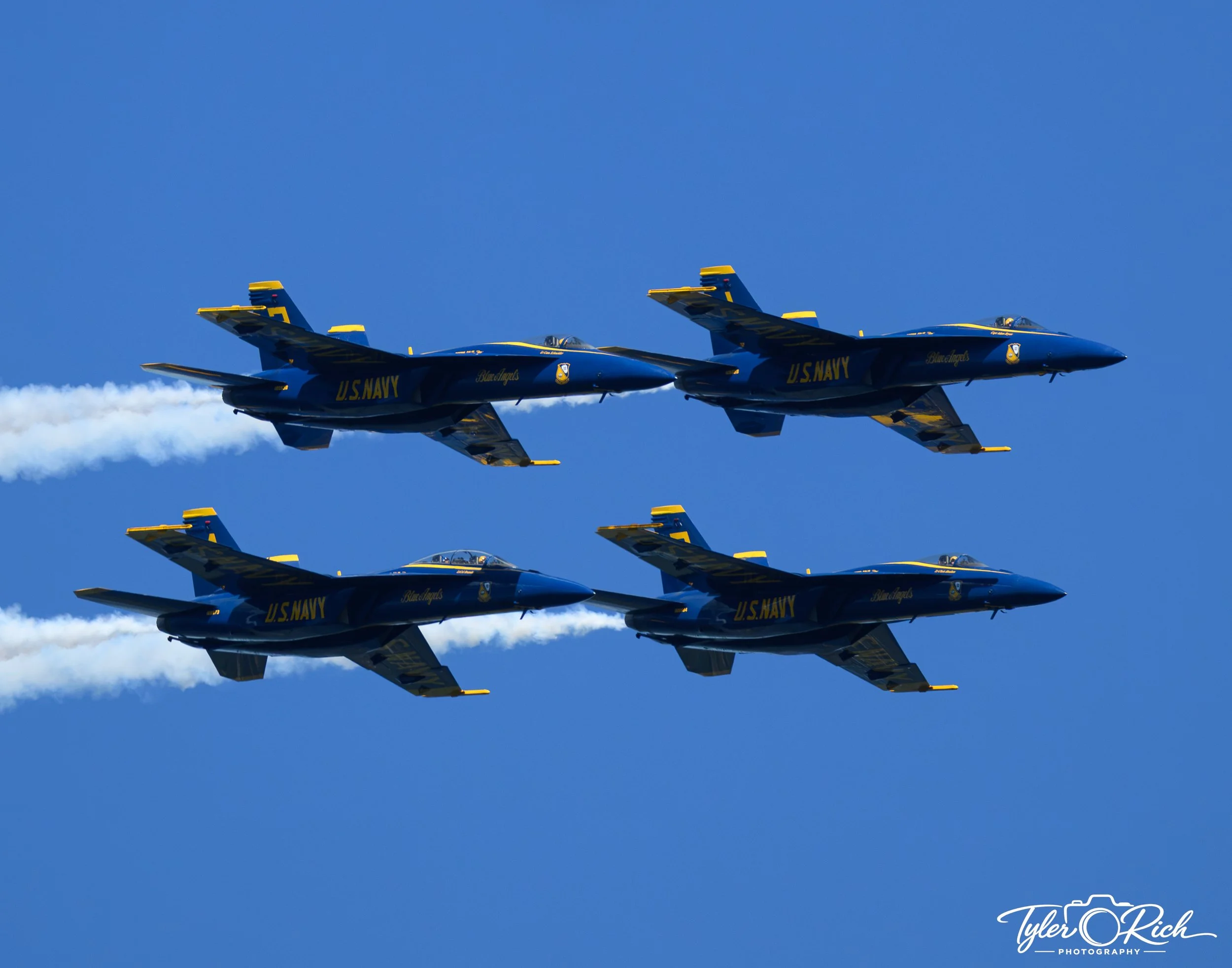 Two U.S. Navy blue and yellow fighter jets flying in formation against a clear blue sky, leaving white smoke trails.