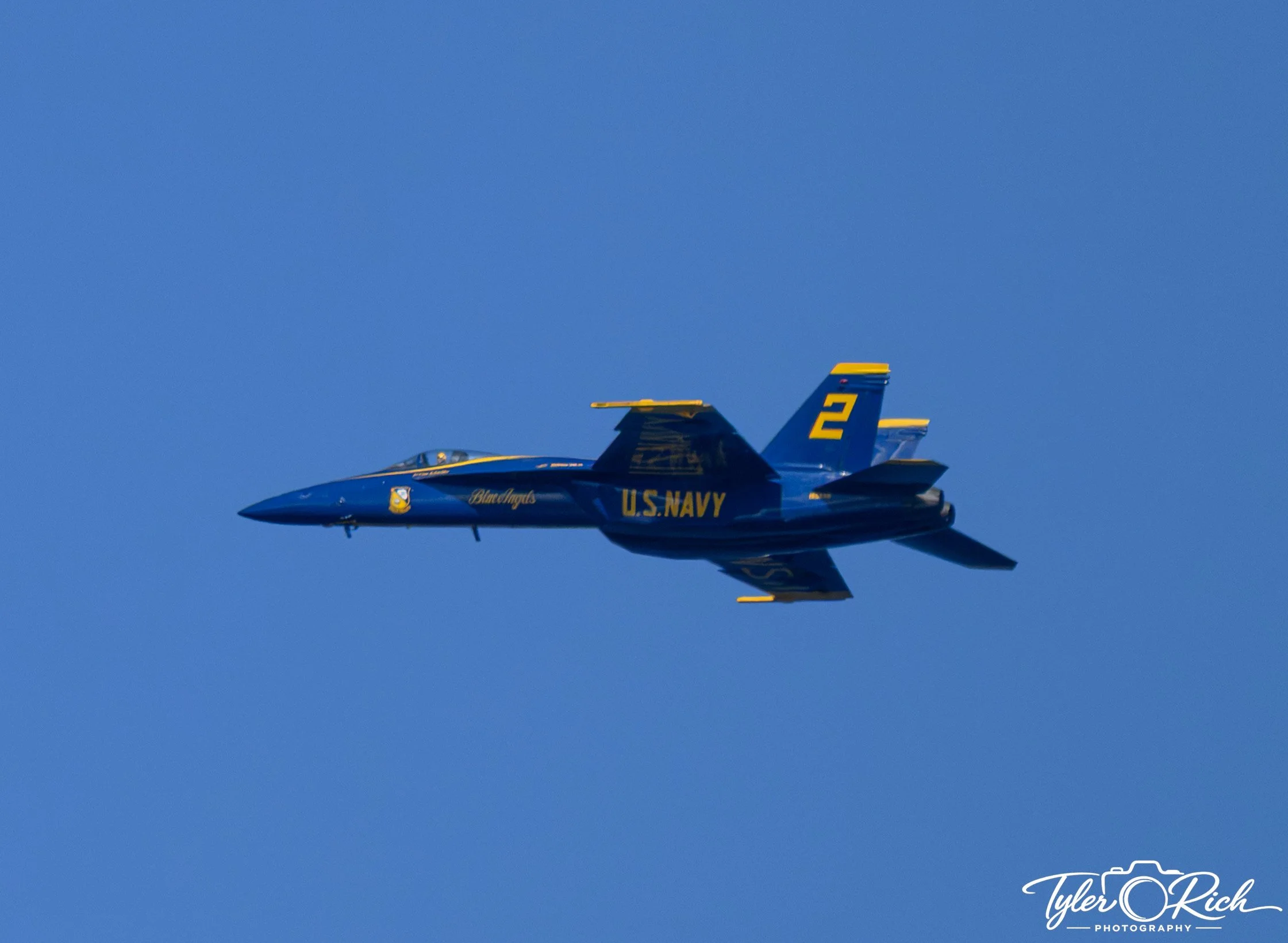 Blue and yellow U.S. Navy fighter jet flying in clear blue sky.