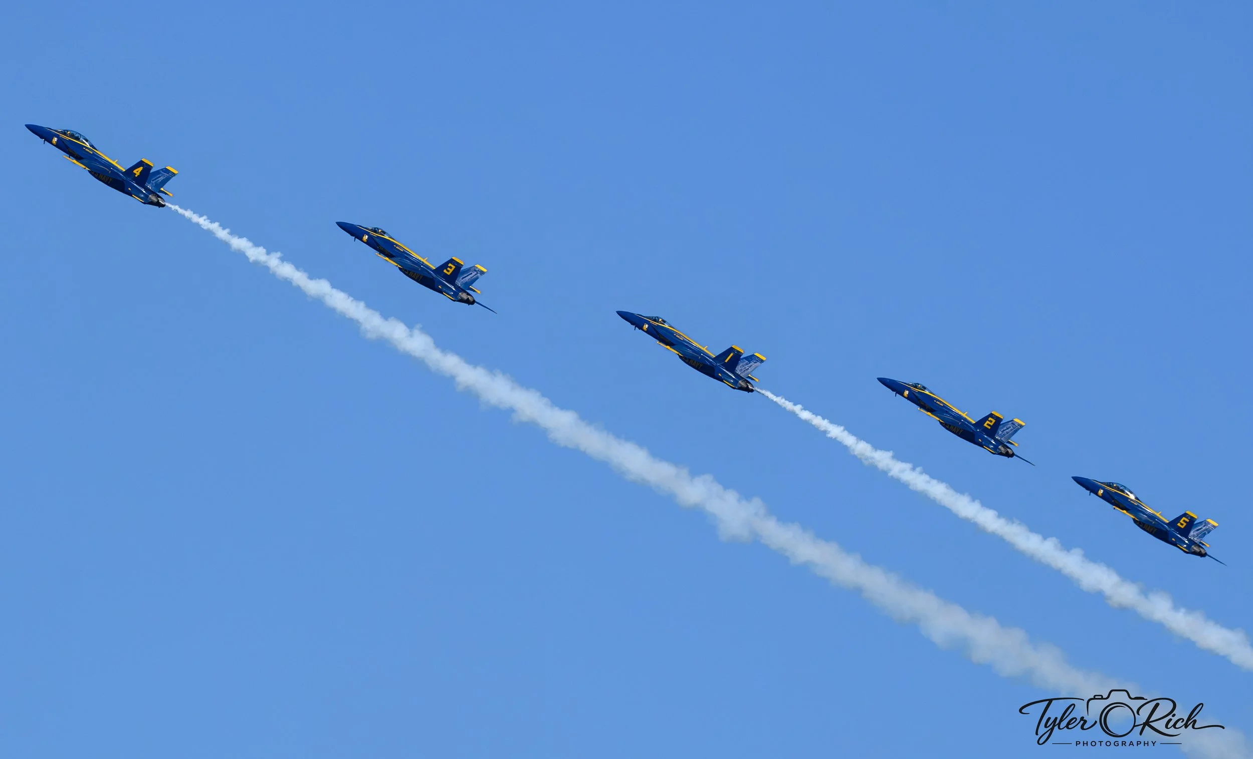 A formation of blue and yellow Navy jets flying in a diagonal pattern in the sky, leaving white smoke trails behind.