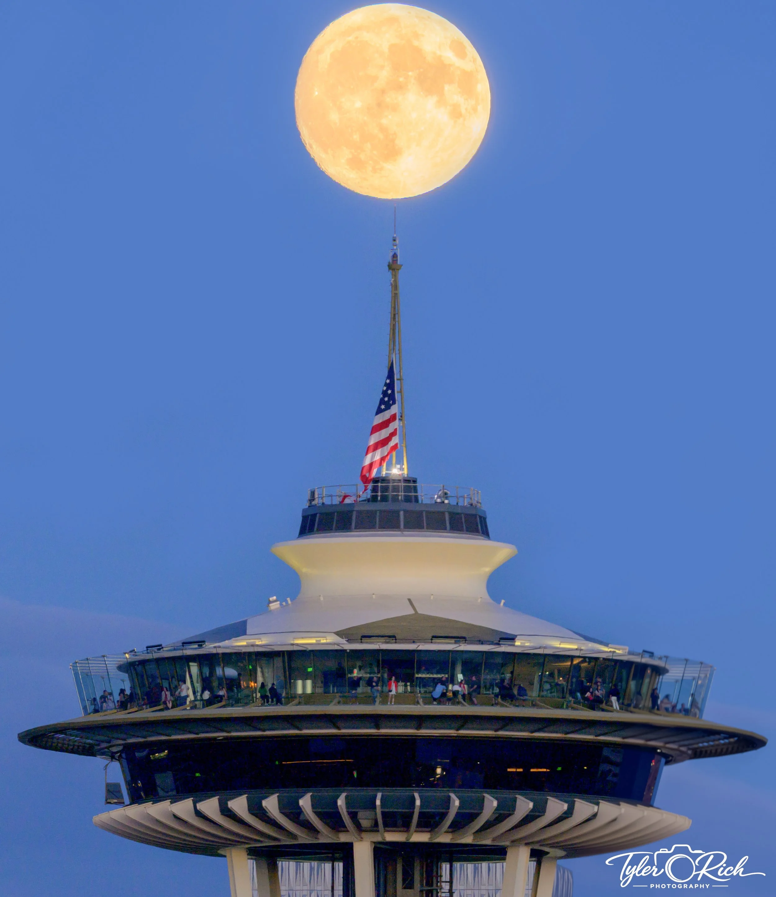 The Space Needle in Seattle during dusk with a large full moon directly above it in the sky, and visitors on the observation deck.