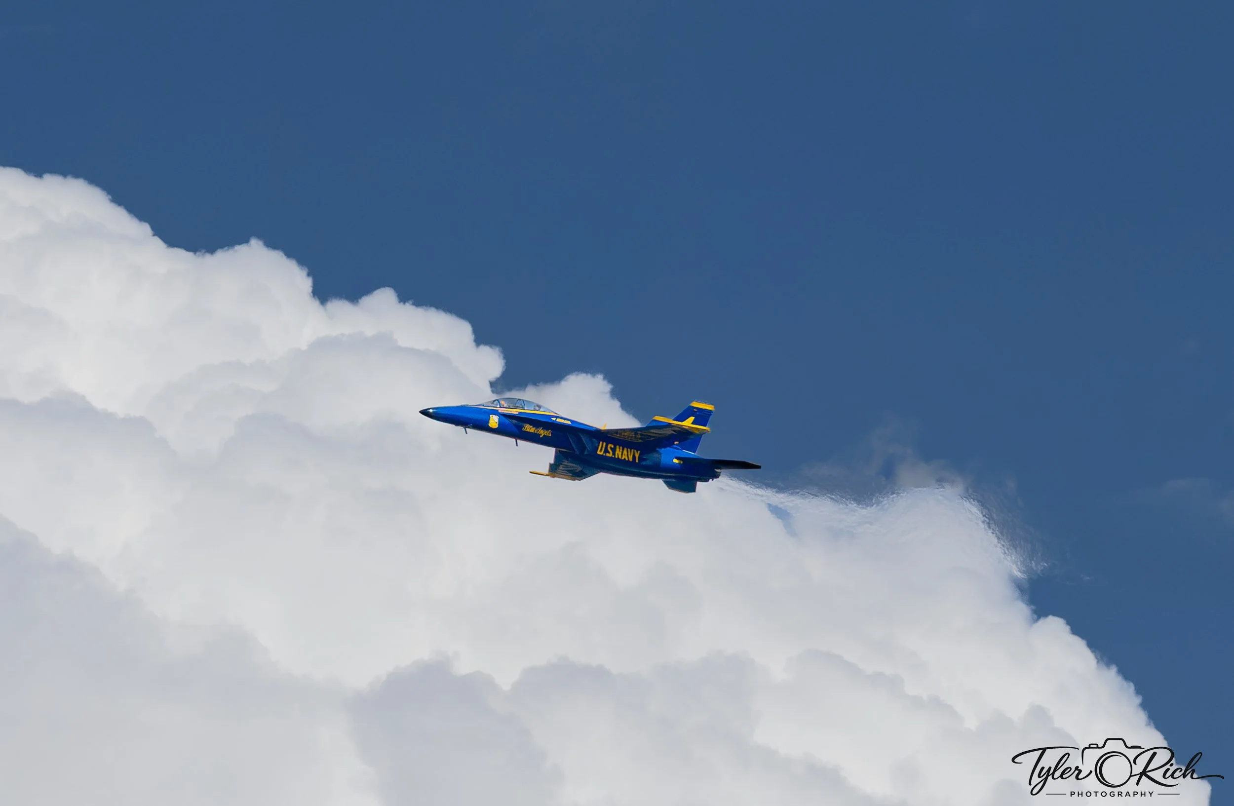 A blue and yellow U.S. Navy fighter jet flying through the sky with white clouds in the background.