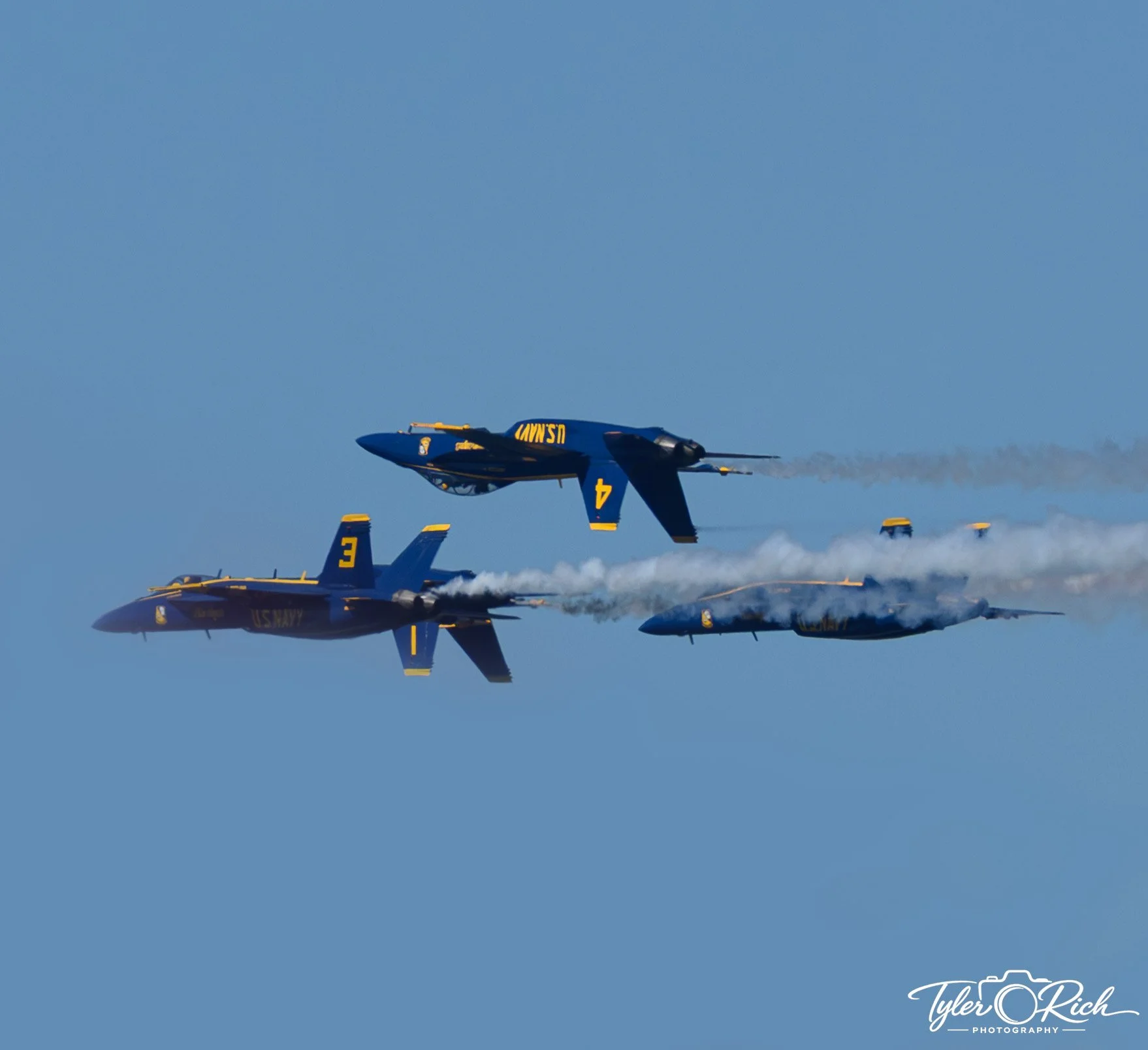 Several US Navy jet planes flying in formation against a blue sky, with trails of smoke behind them.