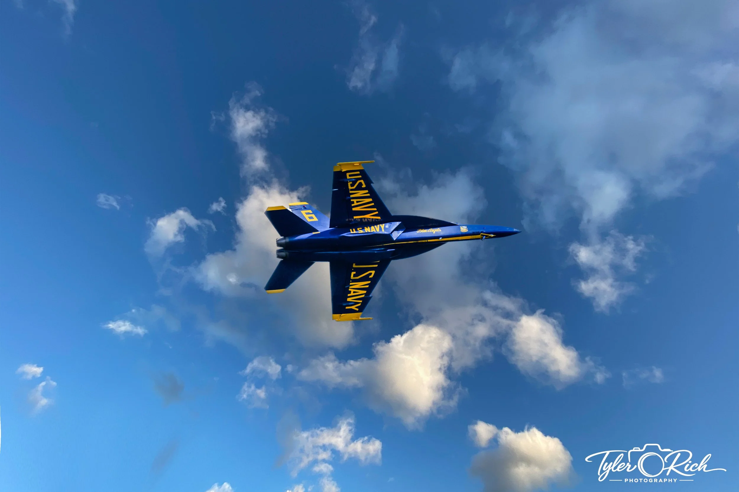 U.S. Navy Blue Angels jet flying through a blue sky with scattered clouds, with the words 'U.S. NAVY' and the number '6' visible on the jet.