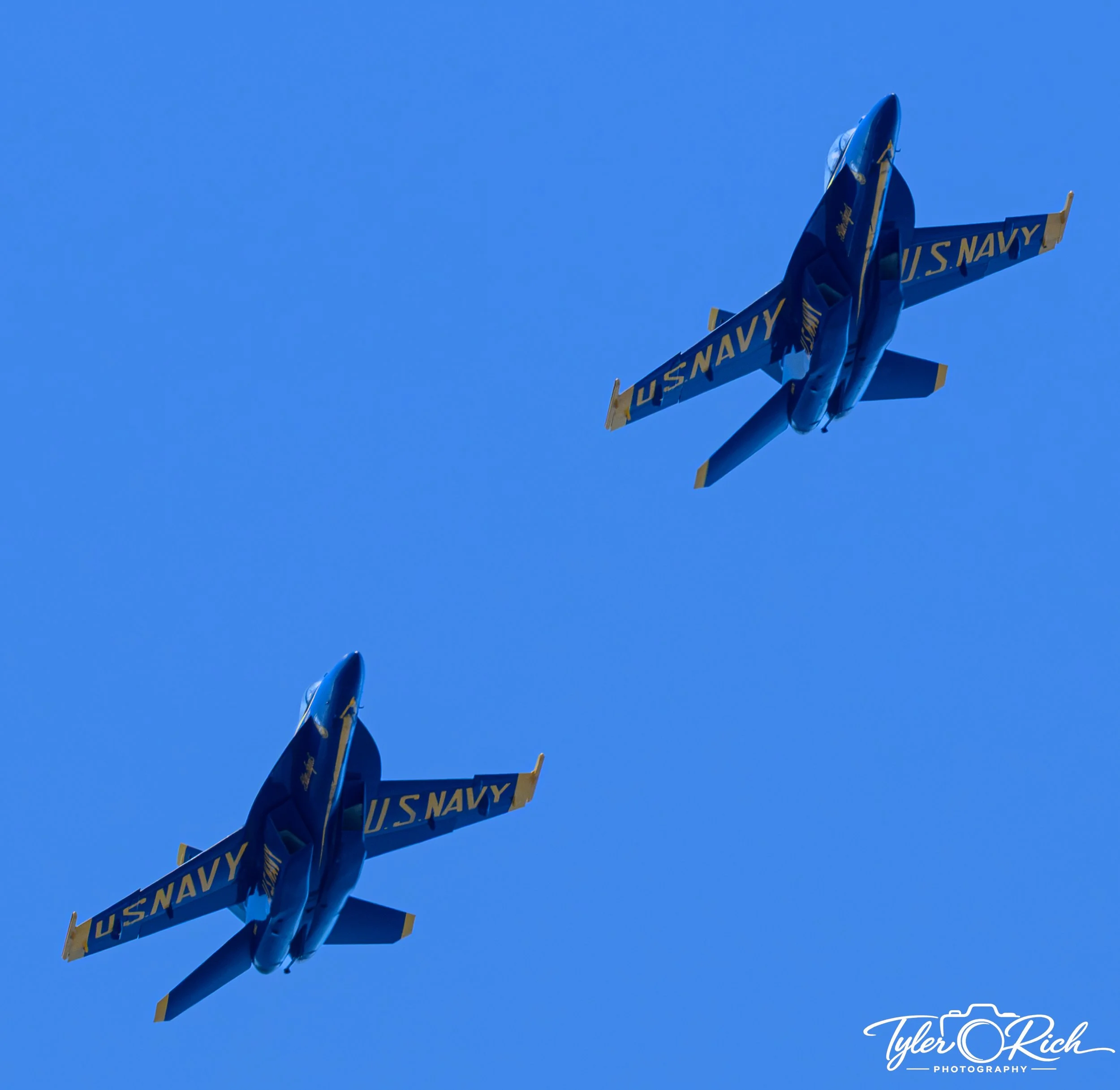 Blue U.S. Navy jet aircraft flying in clear blue sky with yellow accents and 'U.S. NAVY' markings.