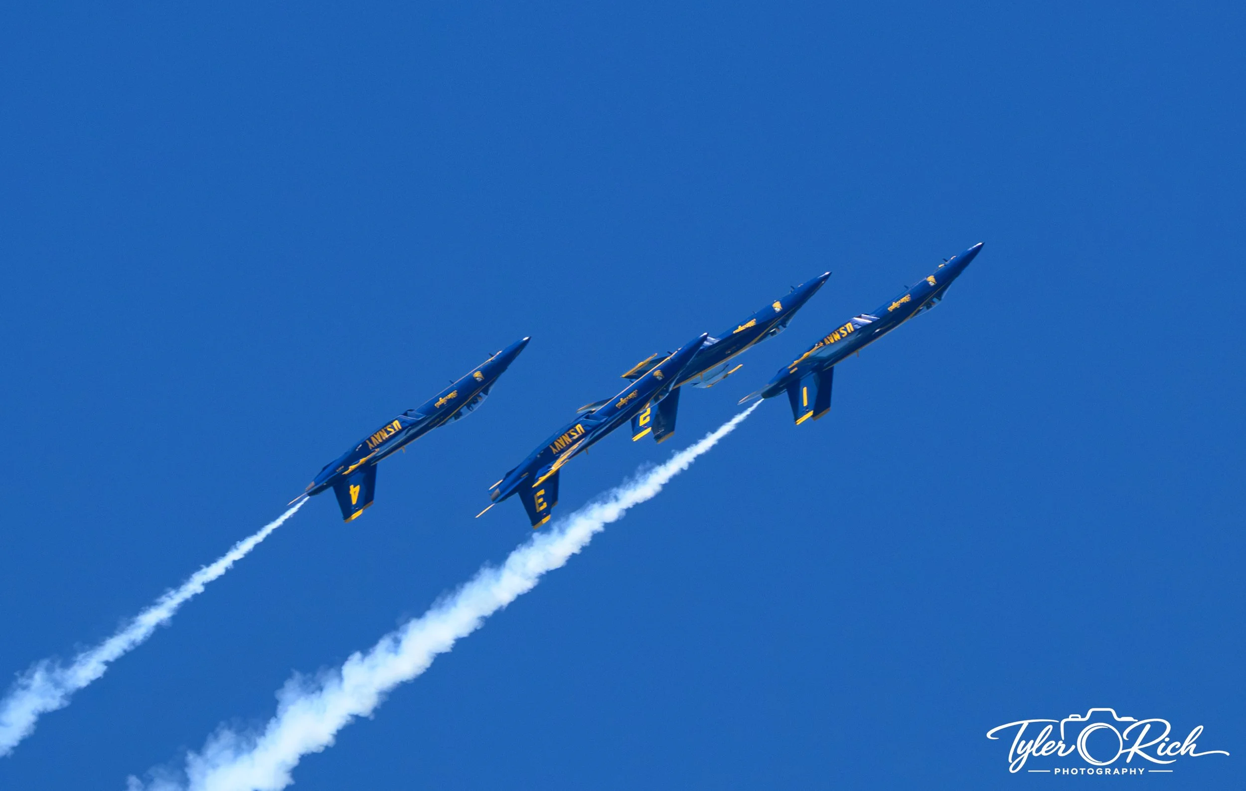 Five blue fighter jets with yellow markings flying in formation against a clear blue sky, leaving white smoke trails behind them.