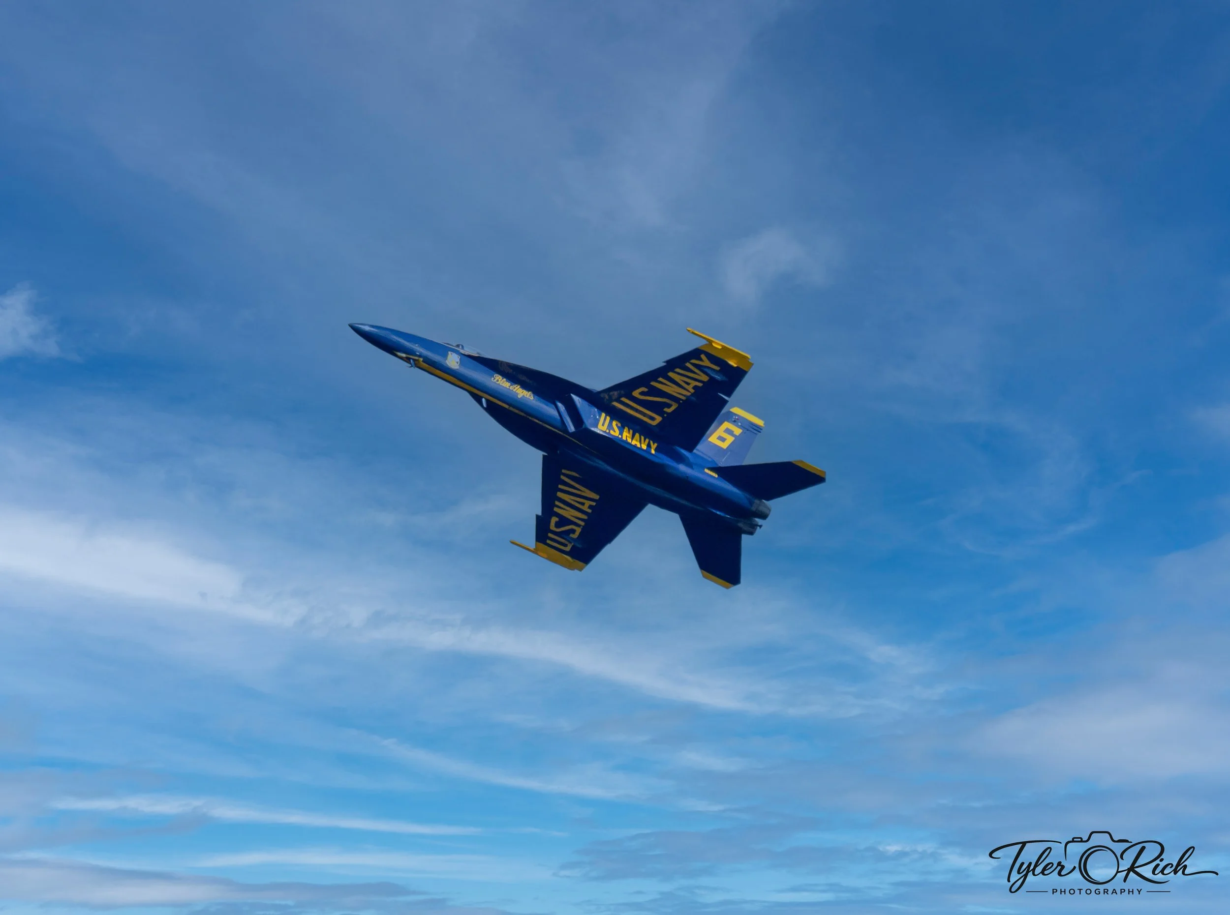 A blue U.S. Navy fighter jet flying in the sky with a trail of white clouds behind it.