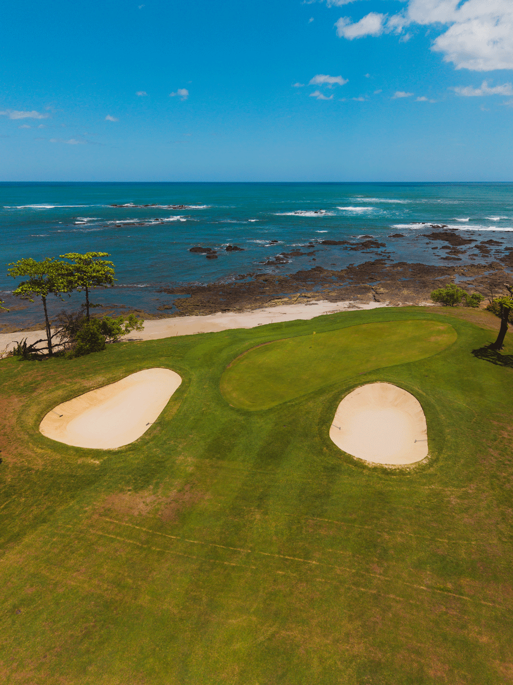 A golf course with green and sand bunkers overlooking the ocean with rocky shoreline and a partly cloudy sky.