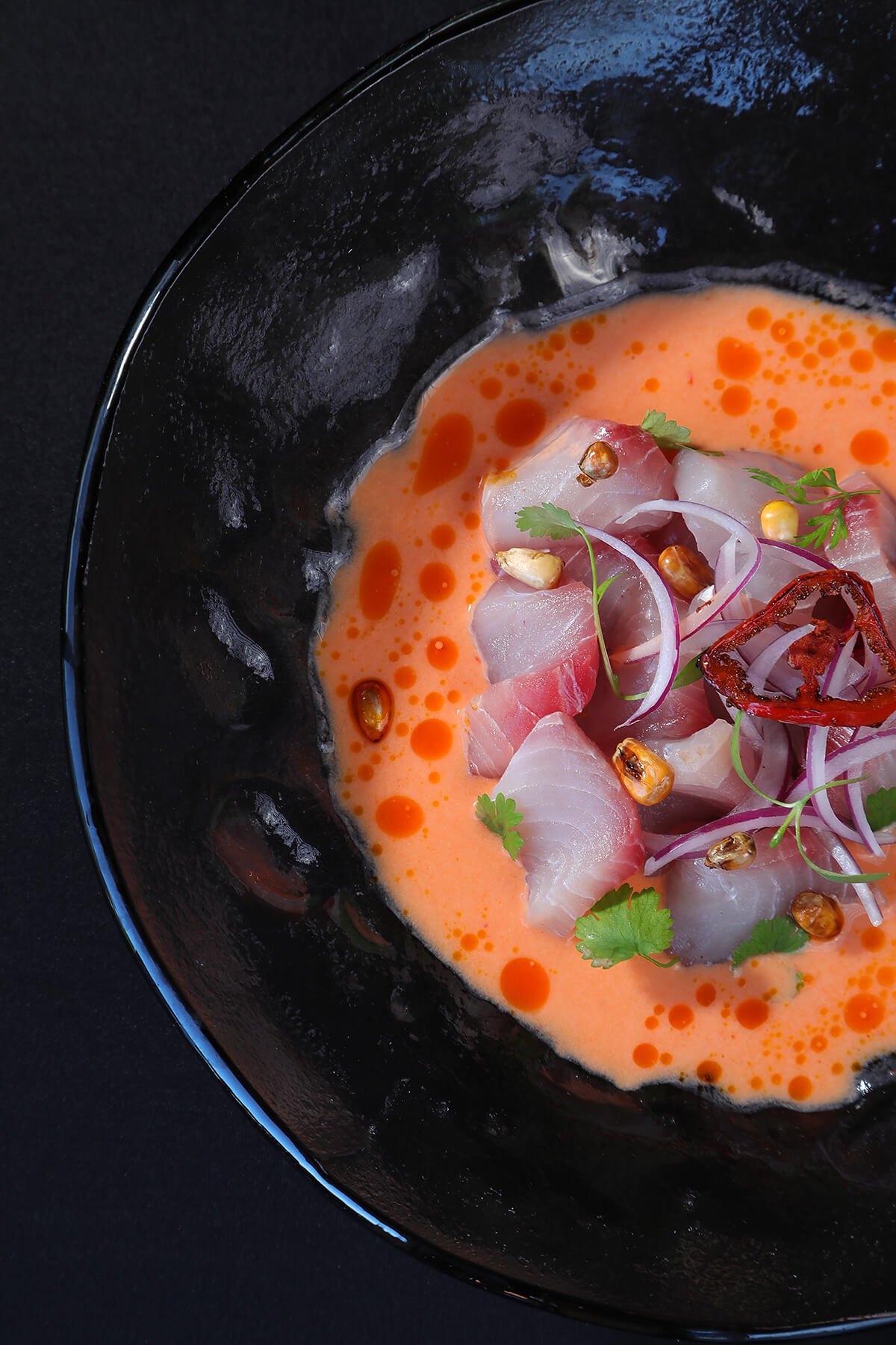 Close-up of a black bowl containing ceviche with pieces of raw fish, red chili, sliced onions, cilantro, corn, and orange sauce.