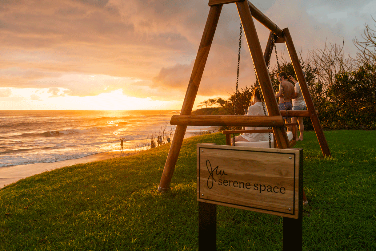 A serene beach scene at sunset with a wooden swing set labeled 'serene space' in the foreground. In the background, people are relaxing and walking along the shore, with the sky painted in warm orange and yellow hues.