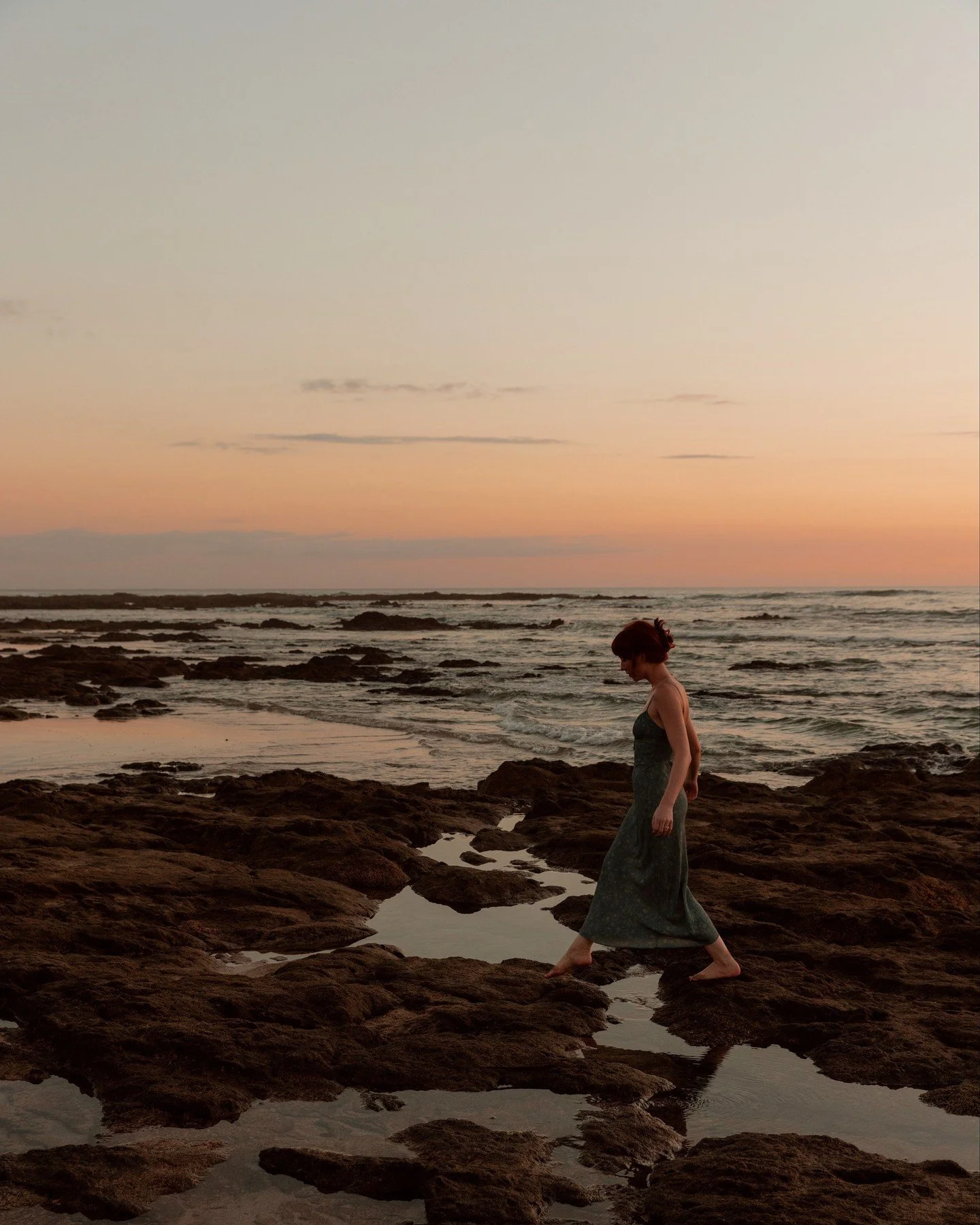 A woman in a dark dress walking barefoot on a rocky shoreline at sunset, with a calm sea and a pastel-colored sky in the background.