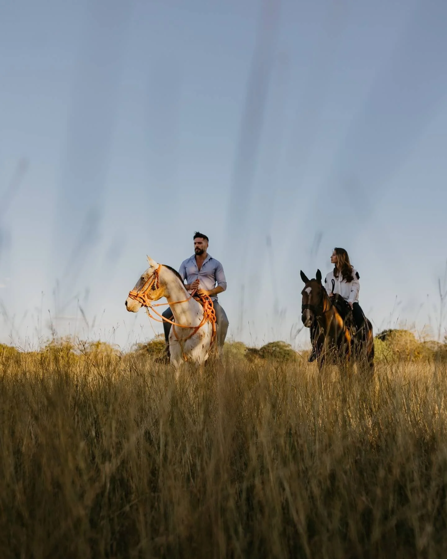 Two people riding horses through a grassy field with a cloudy sky overhead.
