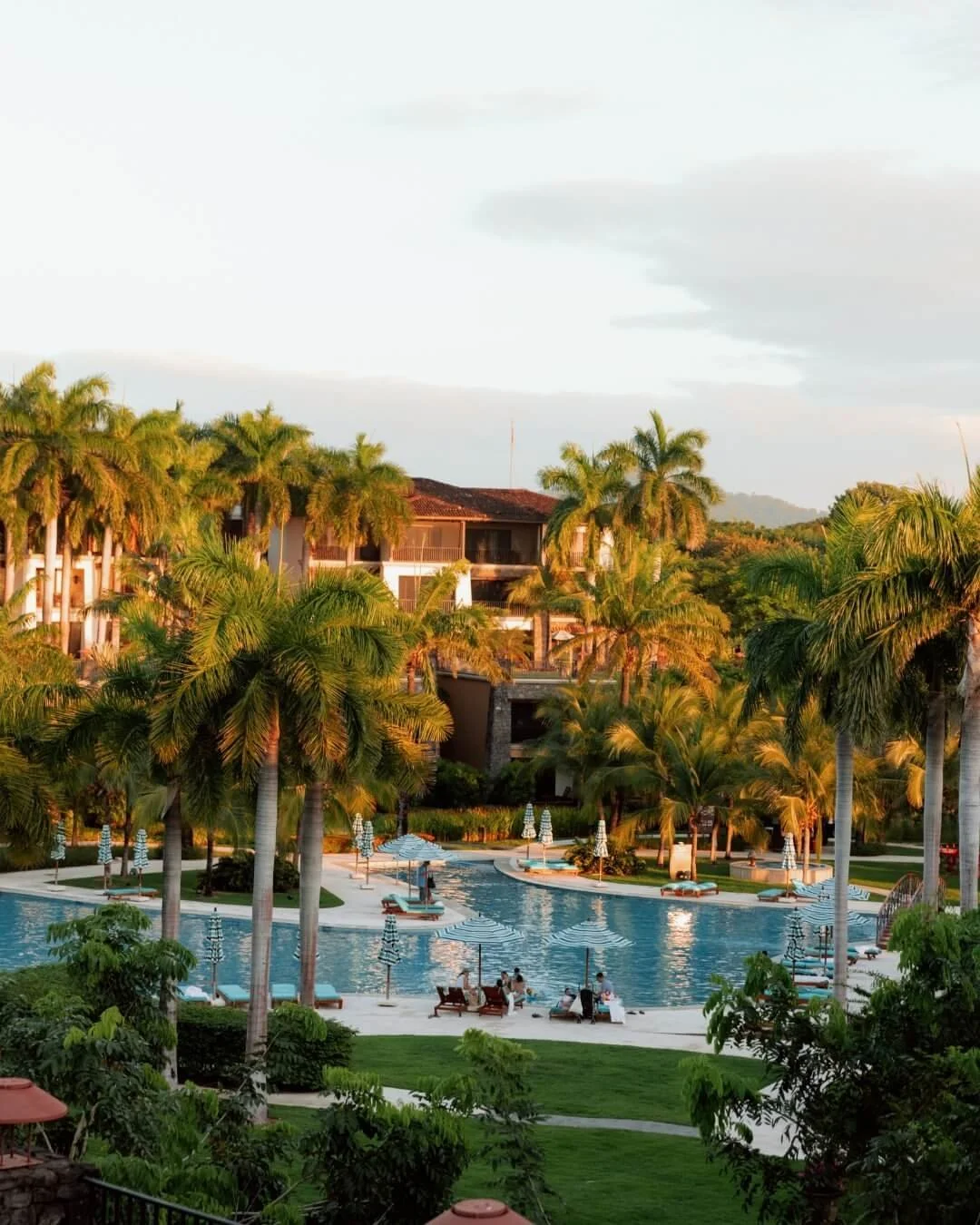 A tropical resort with a large swimming pool surrounded by palm trees, lounge chairs, and umbrellas, with structures in the background and a partly cloudy sky.
