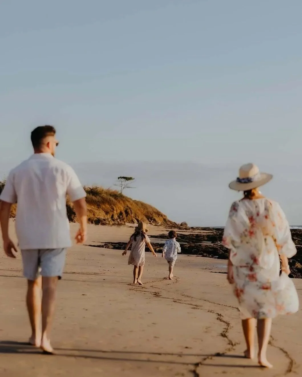 Family walking on the beach during sunset, with a man, woman in a floral dress and hat, and two children running ahead.