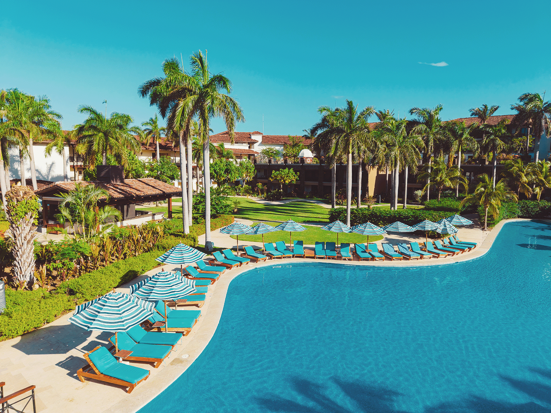 Resort pool area with blue lounge chairs and striped umbrellas, surrounded by palm trees and lush greenery, with buildings and clear blue sky in the background.