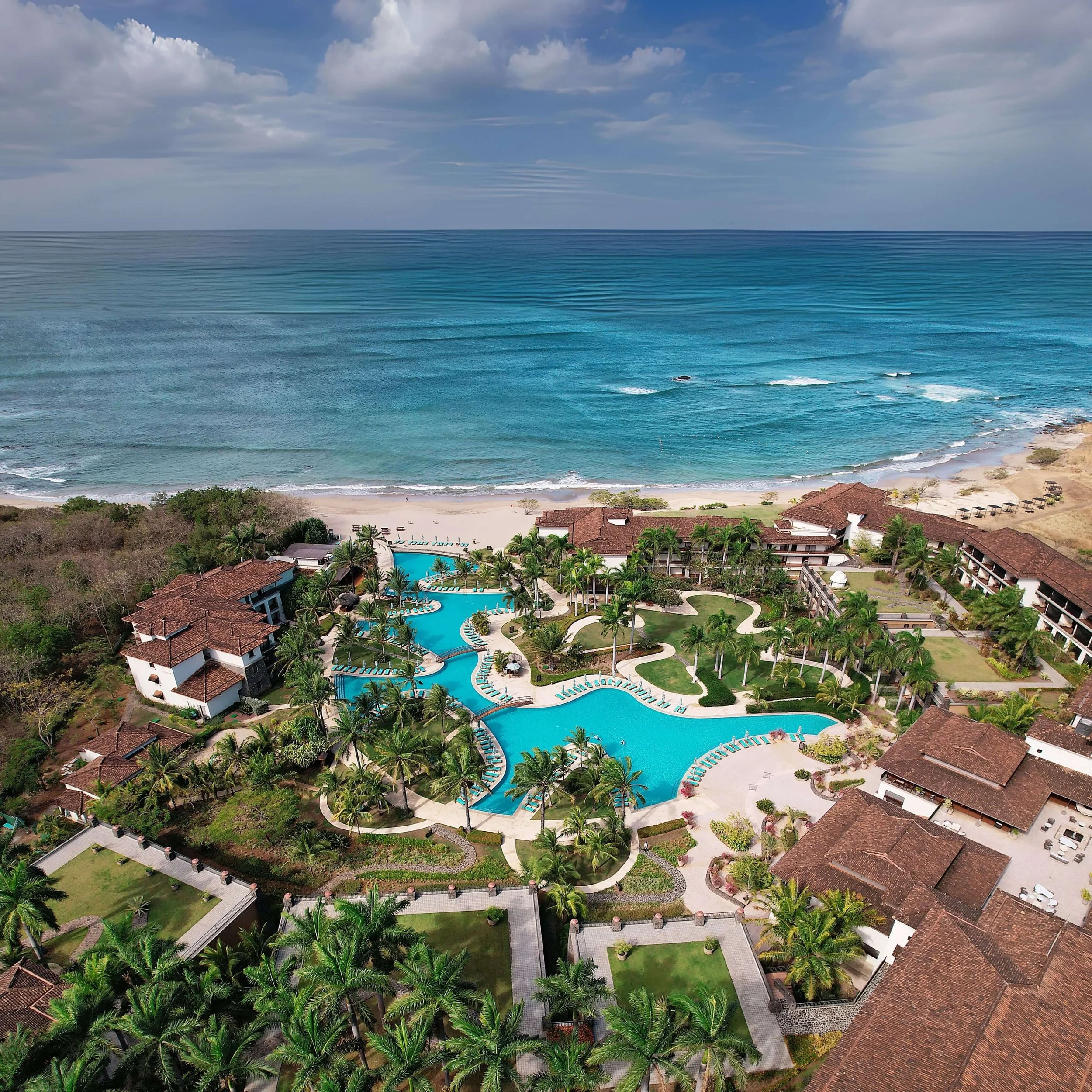 Aerial view of a tropical resort with a large swimming pool, surrounded by palm trees, with buildings featuring red-tile roofs, overlooking the ocean with clear blue water and partly cloudy sky.
