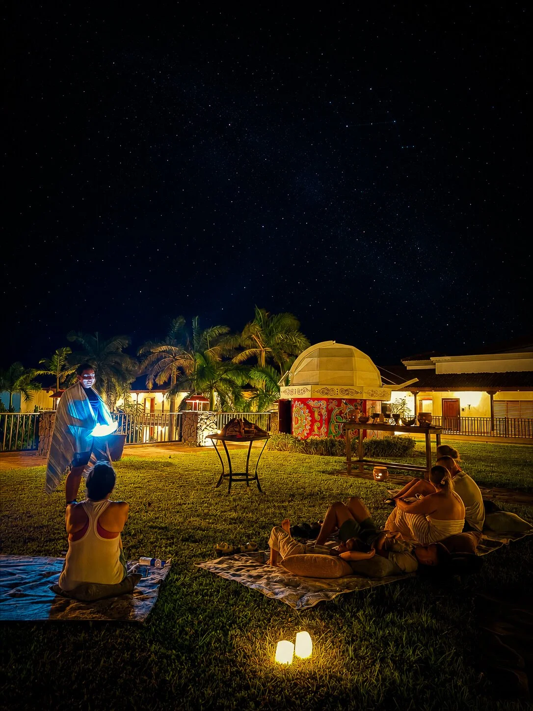 People sitting on blankets and cushions on a grassy area at night, watching a person in traditional attire performing with a glowing object. The scene is illuminated by candles and soft lighting, with stars visible in the sky and palm trees in the background.