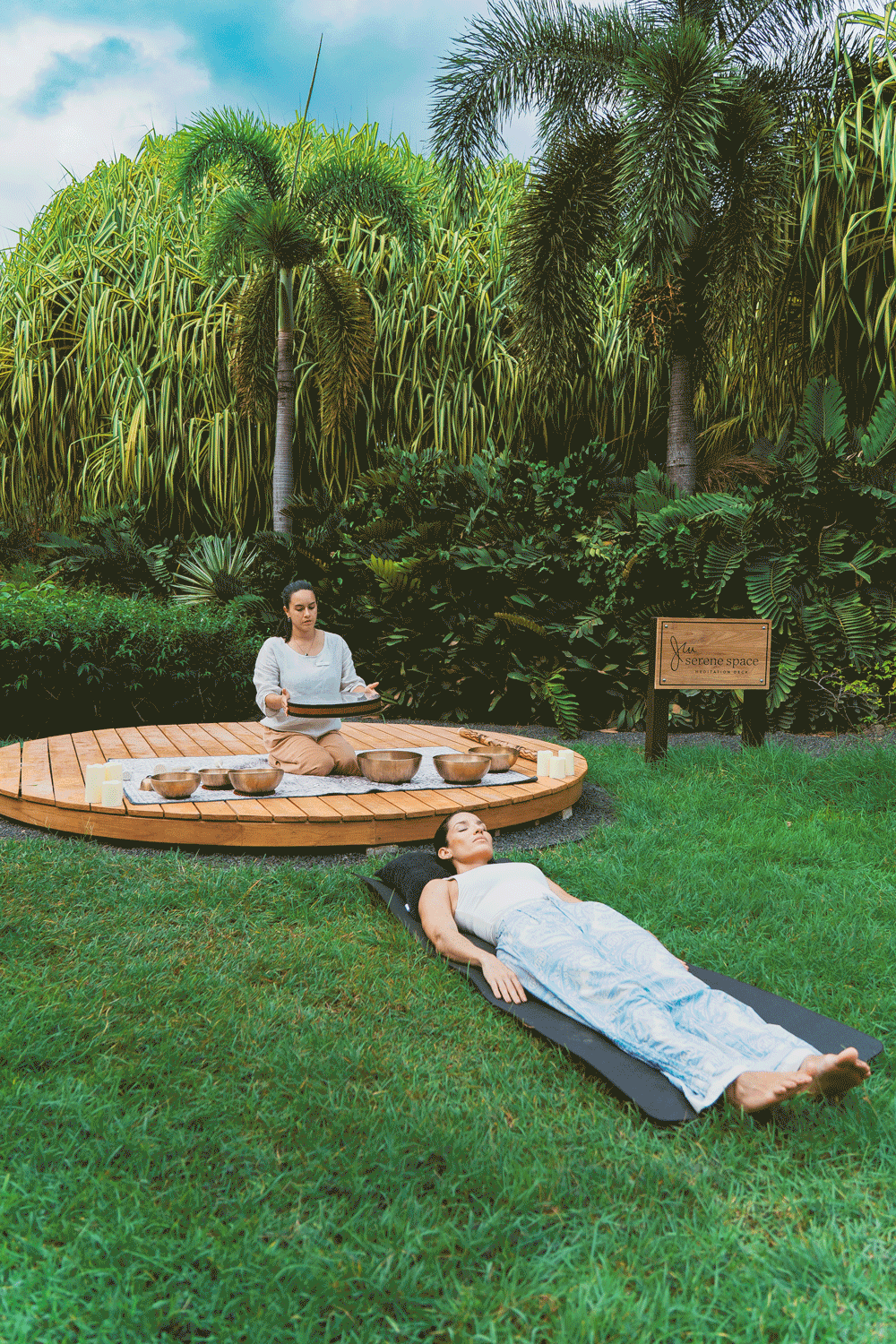 A woman lying on a yoga mat outdoors with lush green plants and trees in the background. Another woman, sitting cross-legged on a circular wooden platform, appears to be playing singing bowls during a meditation or relaxation session in a tranquil garden setting.