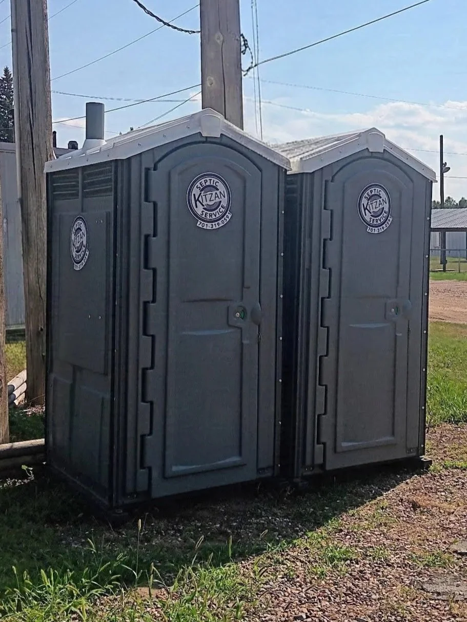 Two portable toilets placed outdoors on a grassy area near a fence and utility pole, with a partly cloudy sky in the background.