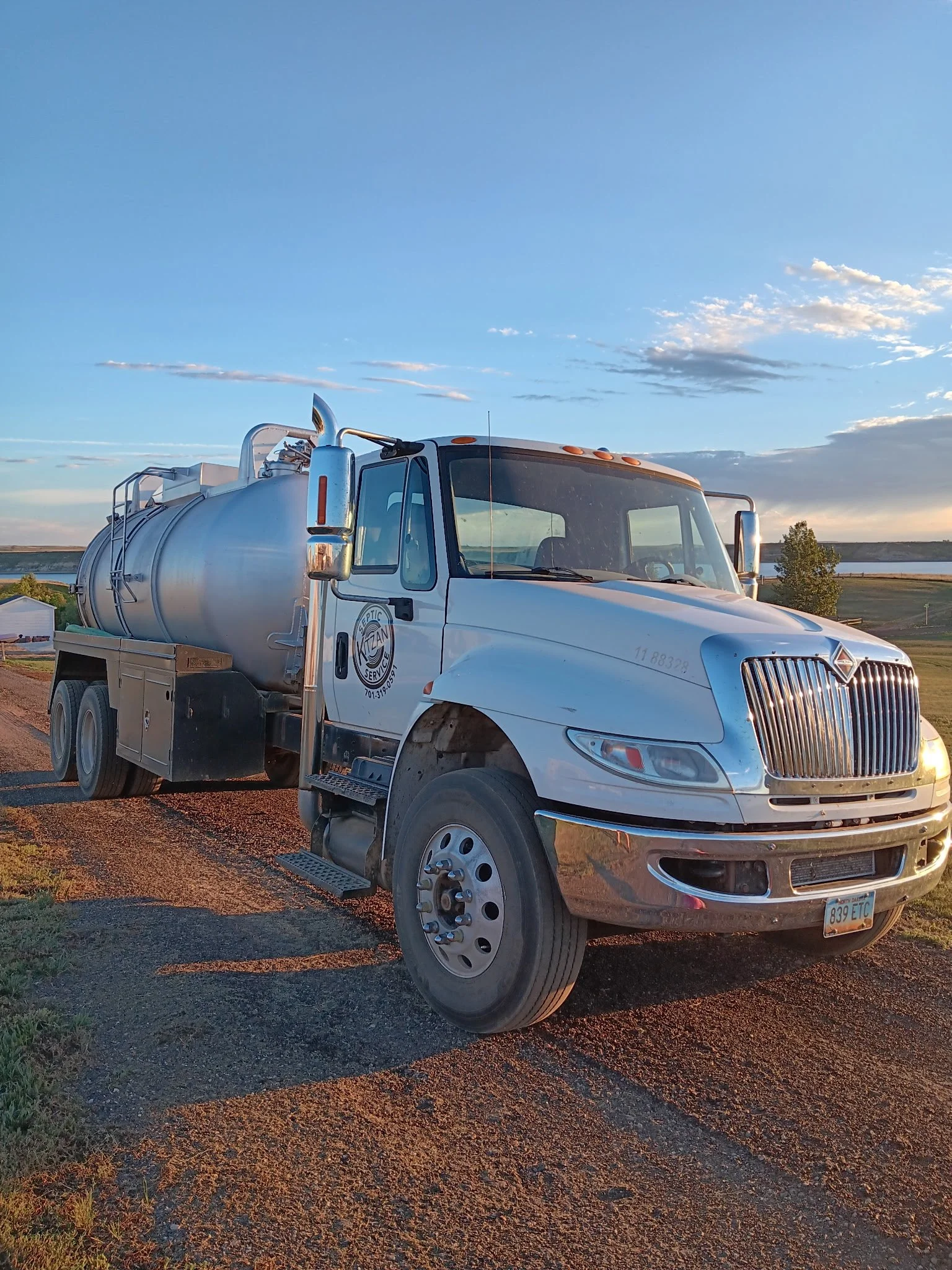 A white tanker truck parked on a dirt road near a body of water with a clear sky and some clouds during sunset.