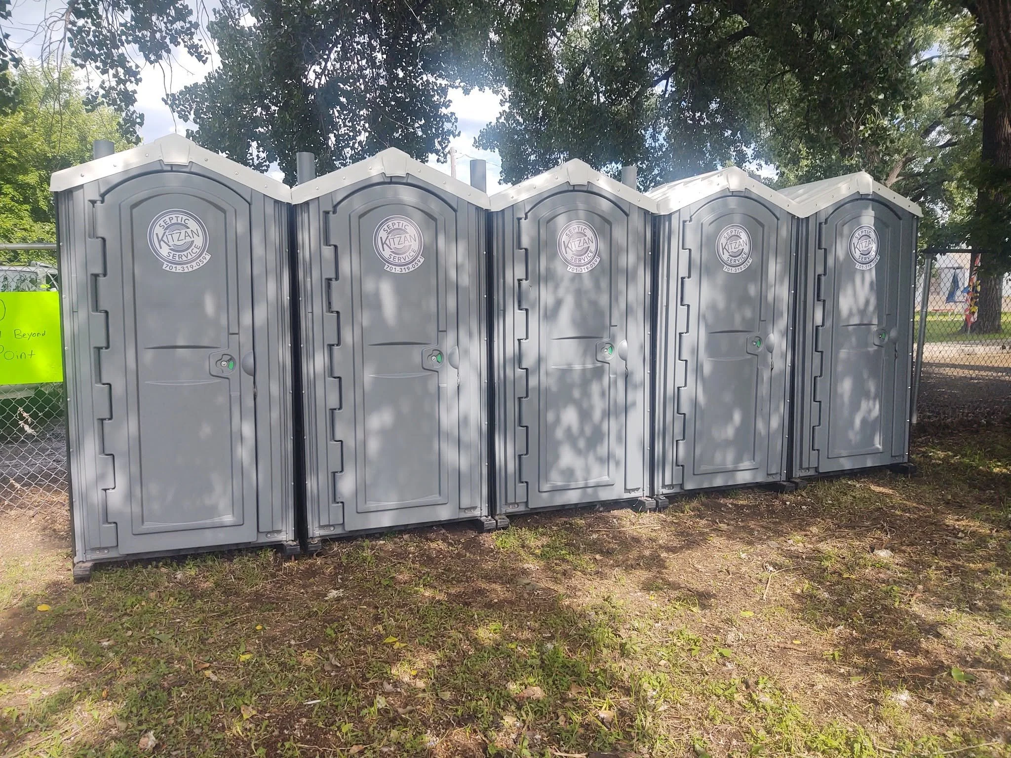 Four grey portable toilets with the logo 'Septic Kitzan Service' and contact number on their doors, positioned outdoors on grassy ground with trees and a chain-link fence in the background.