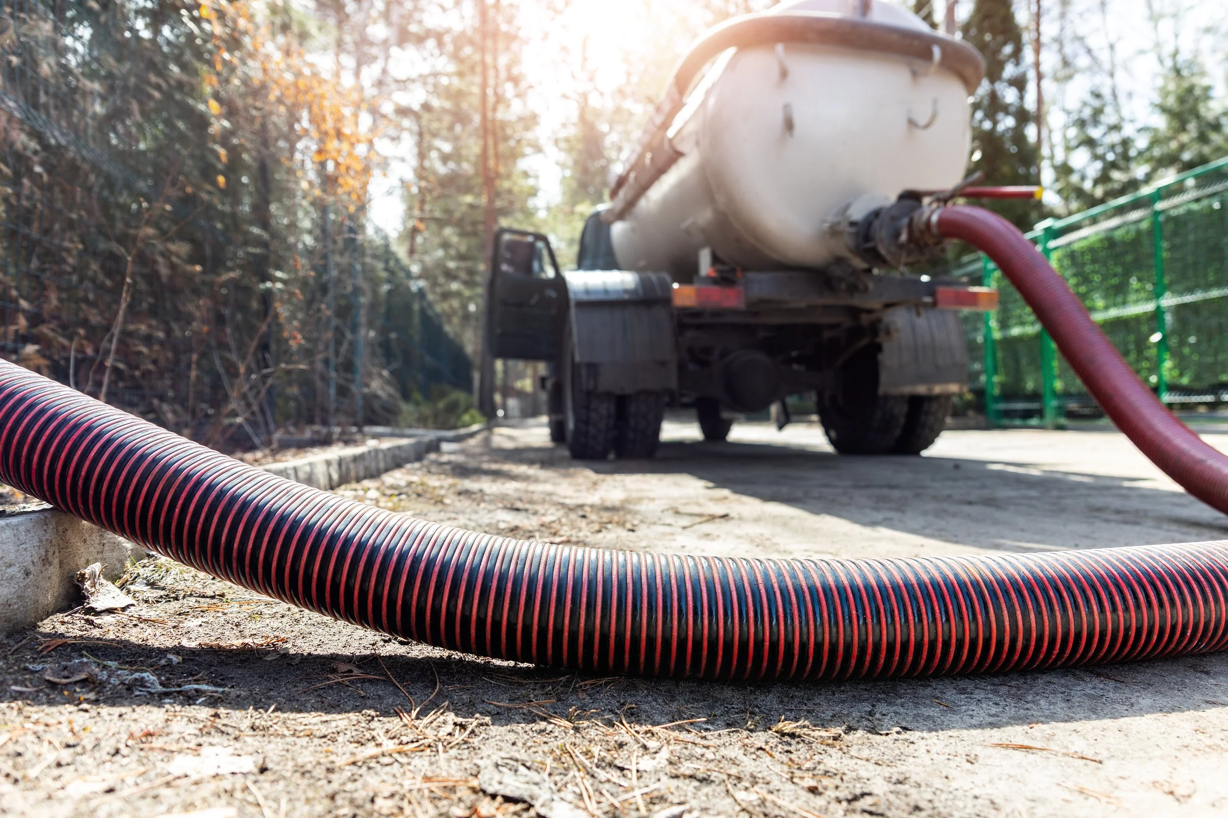 A large truck with a tank is pumping water through a hose onto a dirt ground in a wooded area, with sunlight filtering through trees.