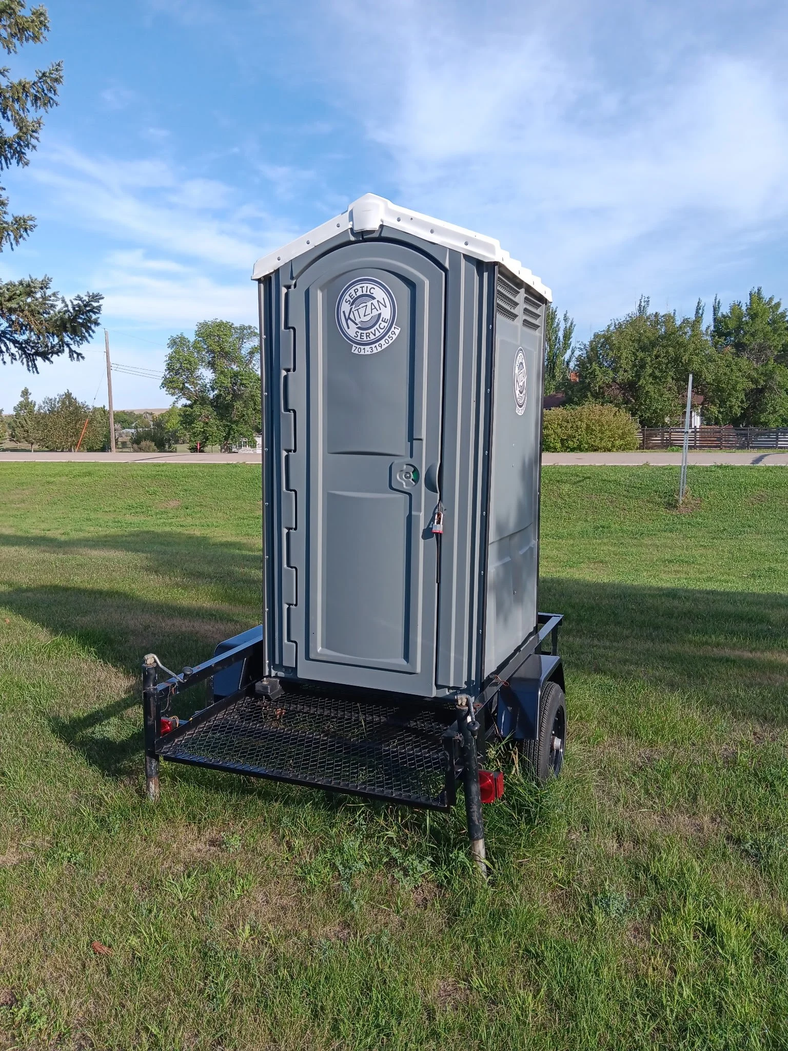 Portable toilet on a black trailer in a grassy park with trees and a blue sky.