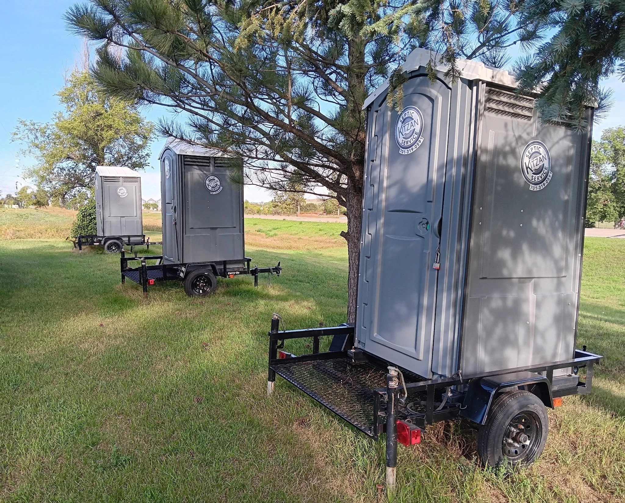 Three portable restroom trailers with gray exteriors and circular logo stickers that say "Septic Kitzan Service" are positioned on a grassy field near trees, with a trailer hitch visible in the foreground and a clear blue sky overhead.