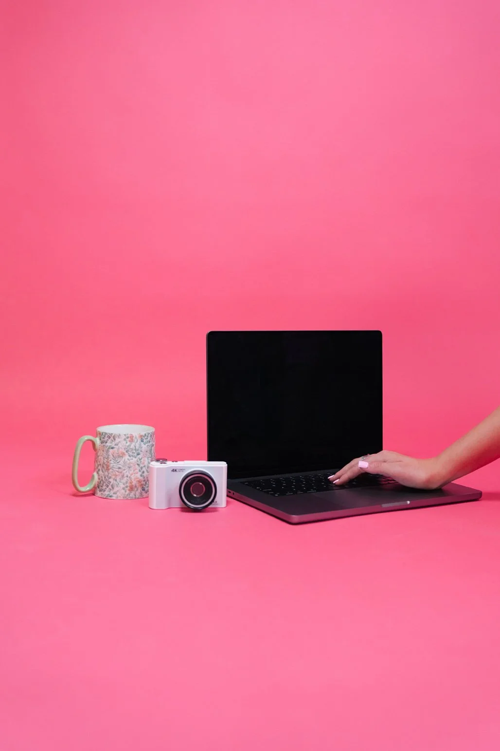 Laptop, white digital camera, and floral mug on a pink backdrop with a hand resting on the keyboard
