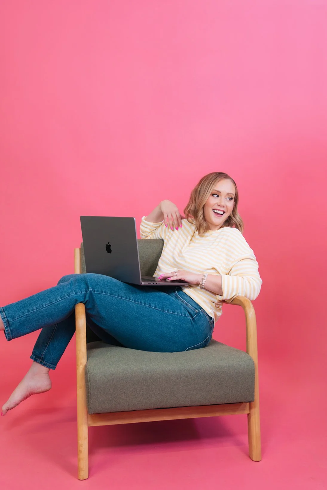 Woman in a yellow striped sweater laughing in a wooden armchair with a laptop on her lap against a pink backdrop
