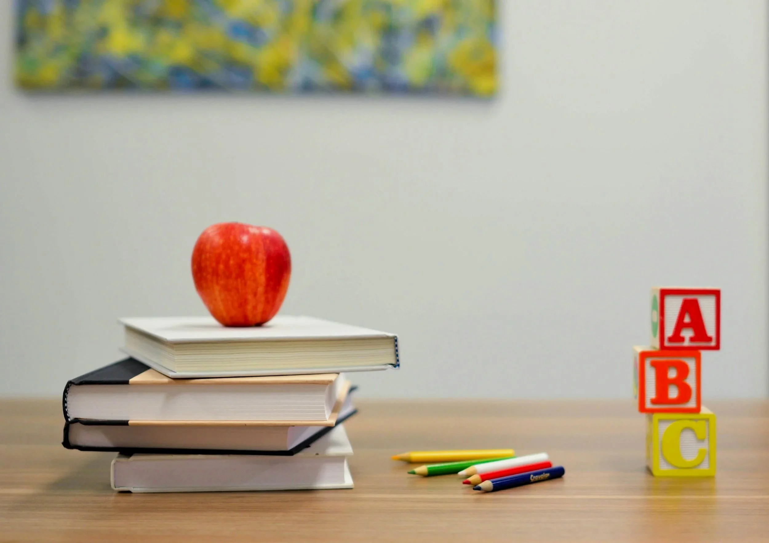 A classroom scene featuring a red apple on top of a stack of notebooks on a desk. To the right, there are colorful alphabet blocks spelling out words and a few colored pencils lying on the desk. A whiteboard with a colorful abstract drawing is in the background.