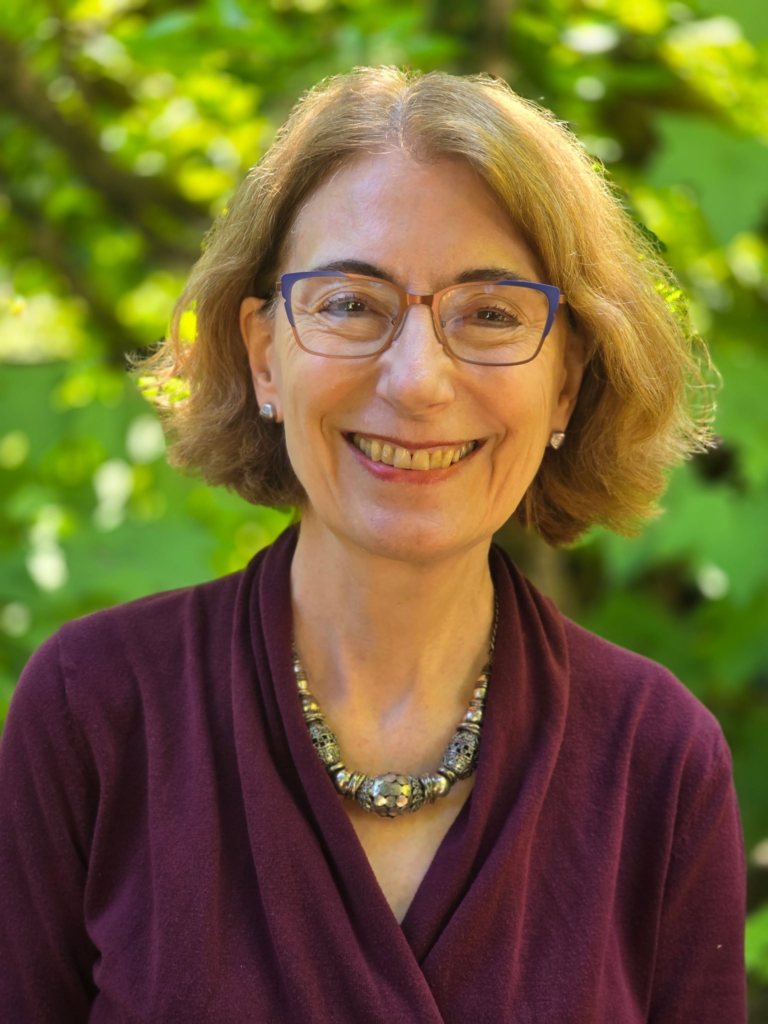 Robin Behn Counselor pictured here with shoulder-length light brown hair, wearing glasses, a dark red blazer, and a statement necklace, smiling outdoors in Oregon with green foliage in the background.