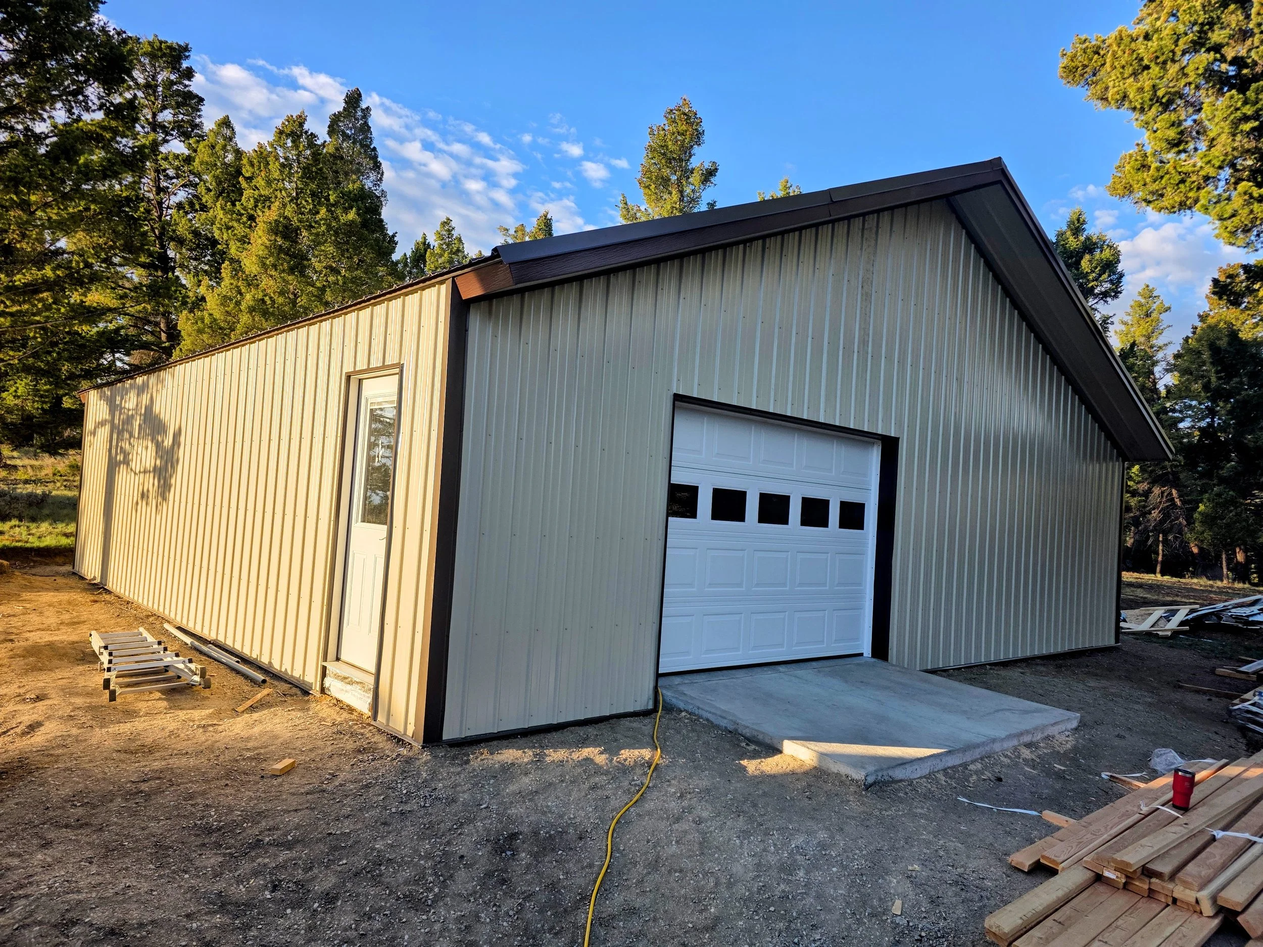 A metal building with a garage door, under construction, with trees in the background and construction materials and tools on the ground.