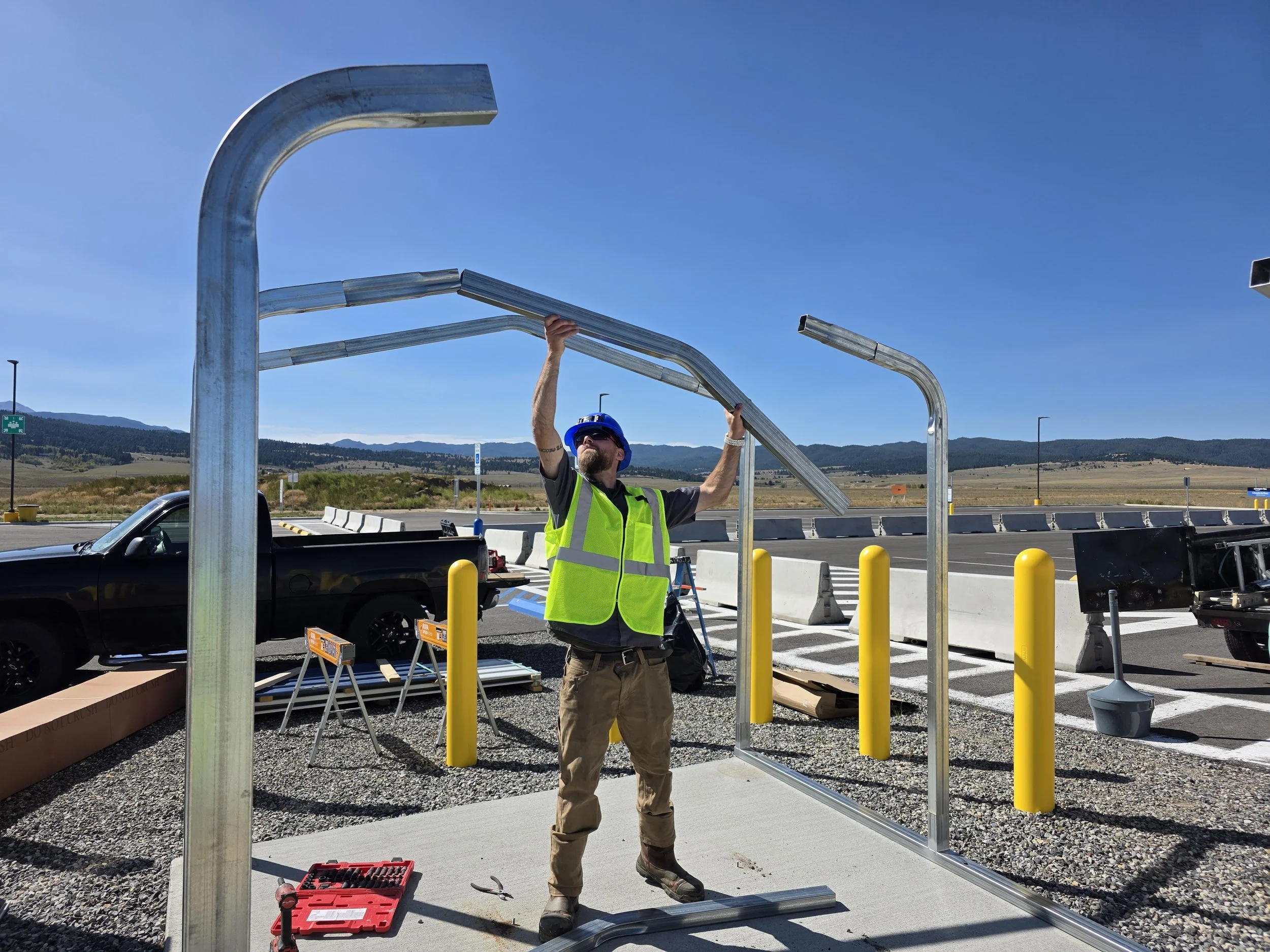 A construction worker wearing a blue helmet and safety vest is installing metal tubing at an outdoor construction site. The background features a clear blue sky, distant mountains, a highway with yellow bollards, and parked cars.