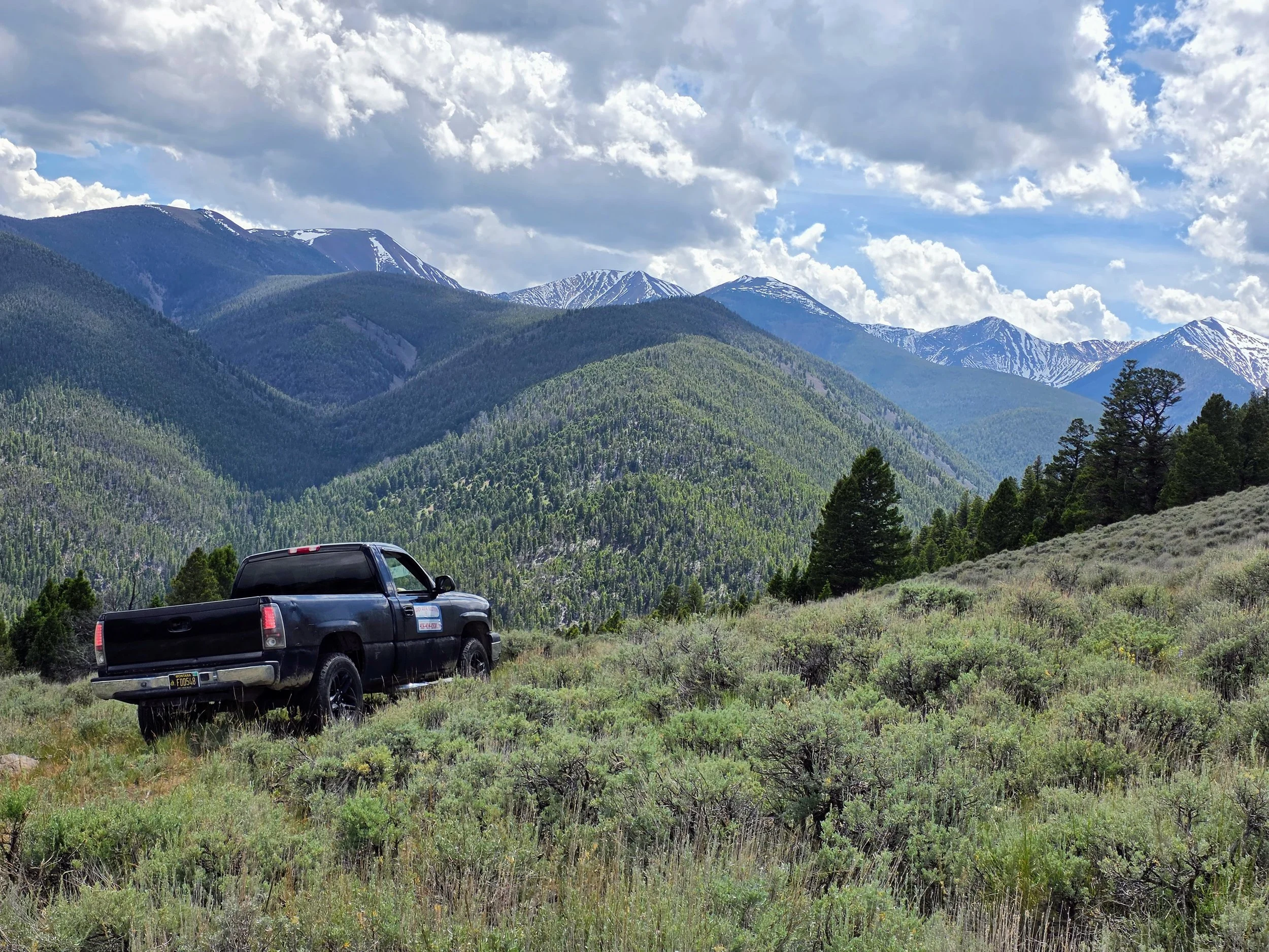 Black pickup truck parked on grassy field with forested mountains and partly cloudy sky in the background in Pipestone, Montana.