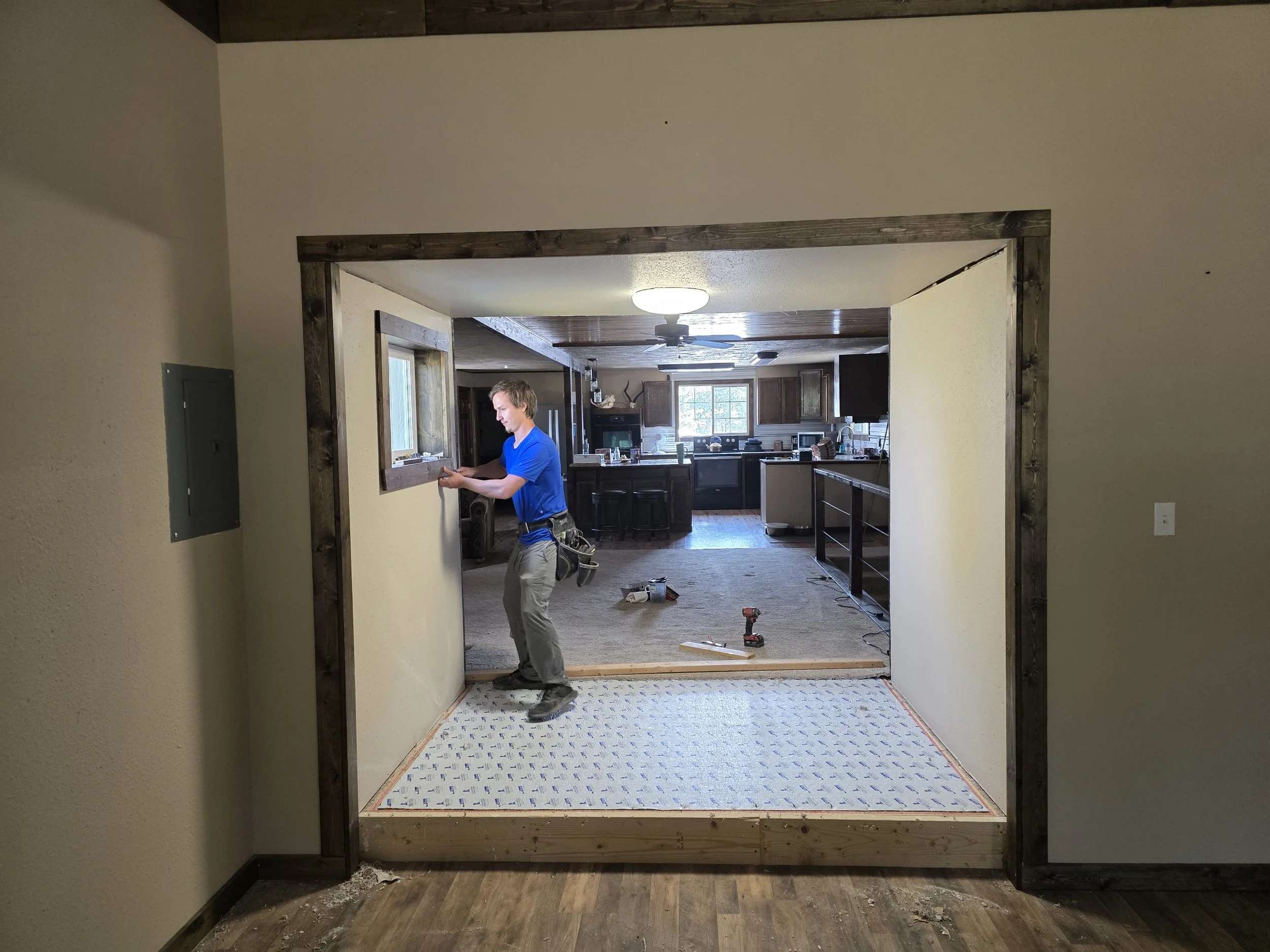 A man in a blue shirt and gray pants working on interior home renovation, standing in a doorway framing a room, with tools on the floor and a kitchen visible in the background.