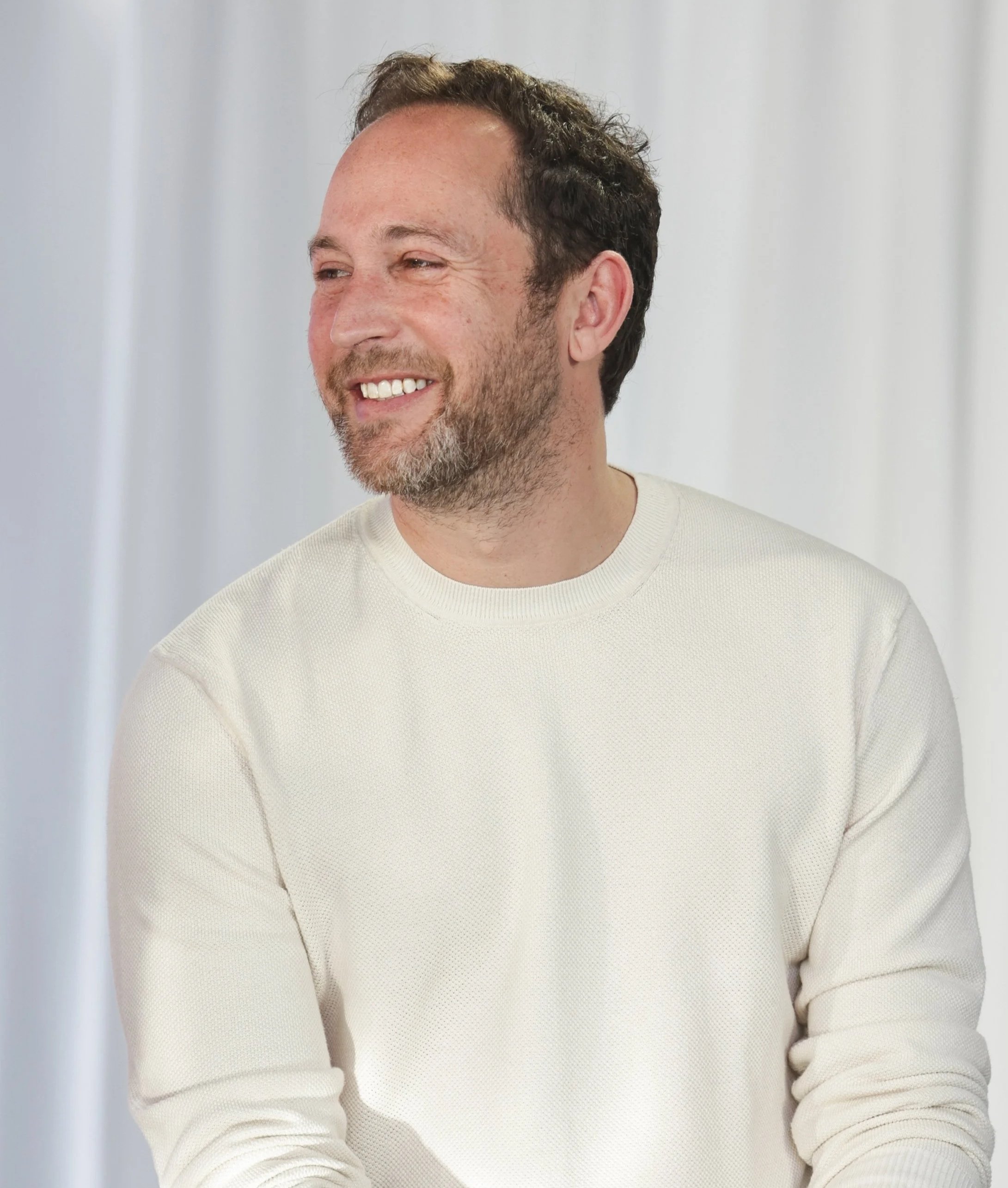 Close-up of a smiling man with short brown hair, beard, wearing a beige sweater, against a light background.