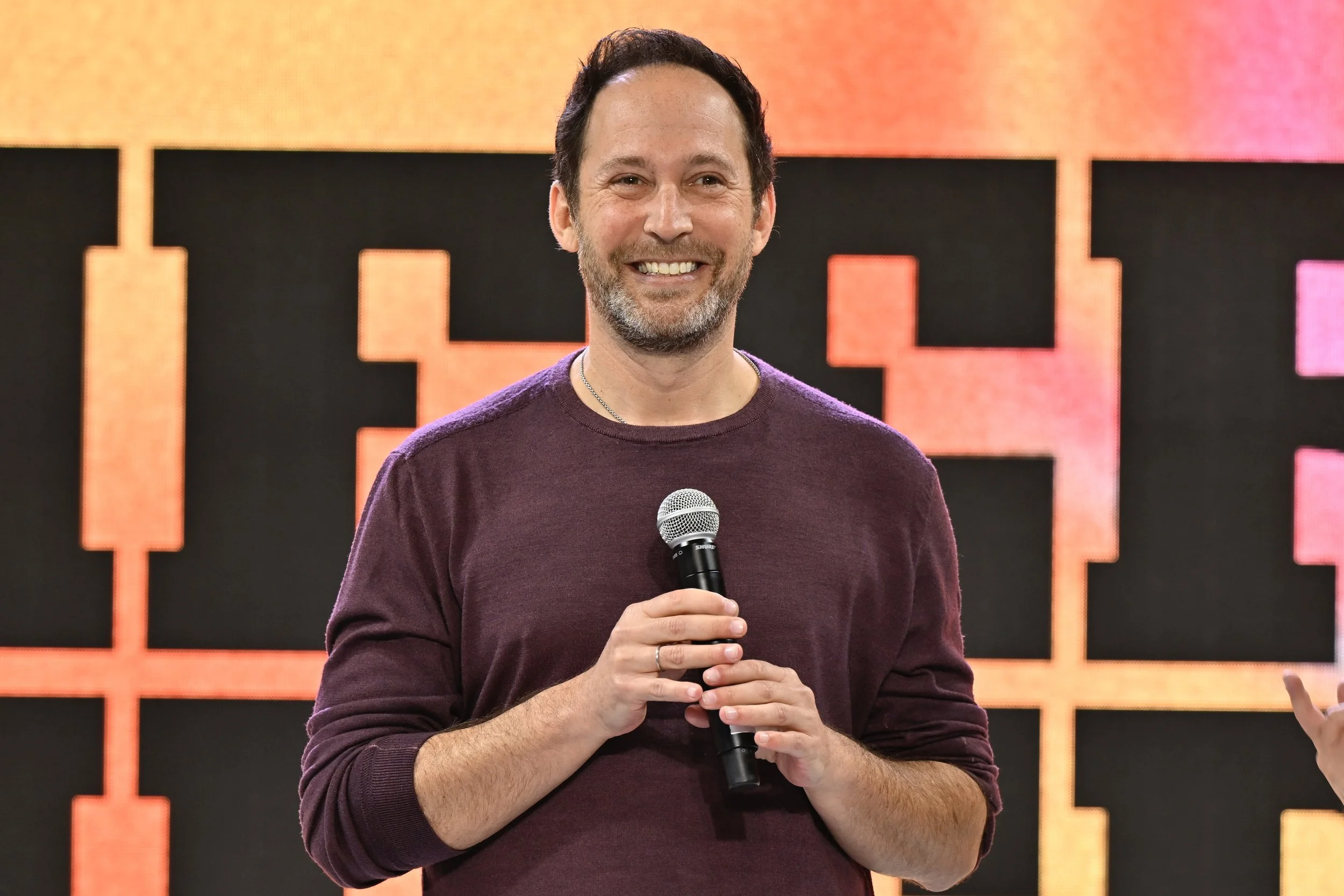A man with dark hair and a beard smiling while holding a microphone.