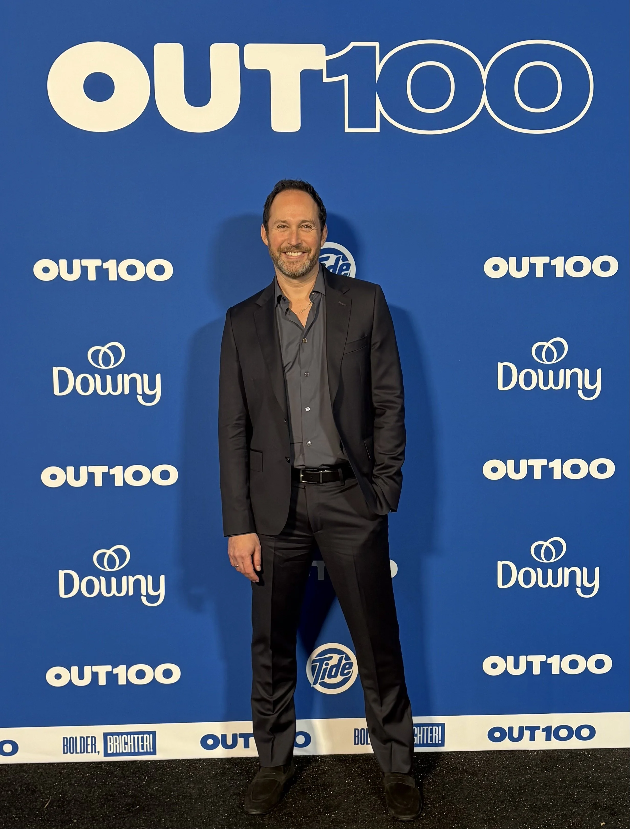 A man in a black suit with a gray shirt standing in front of a blue backdrop featuring the 'OUT100' logo.. The man is smiling with a beard and short dark hair.