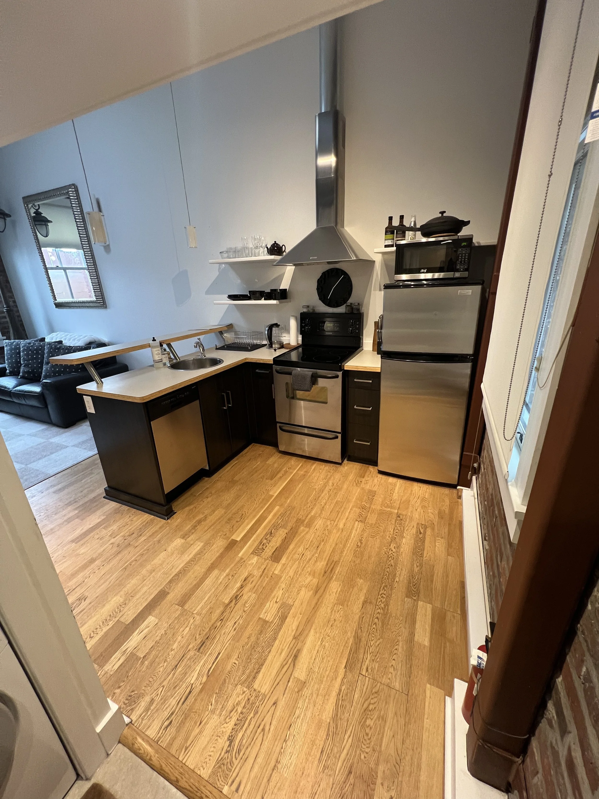 View of a kitchen area with wooden flooring, black cabinets, a stainless steel refrigerator, microwave oven on top, and a stove with an overhead stainless steel range hood. To the left, a countertop with a sink, dish drying rack, kettle, napkins, and