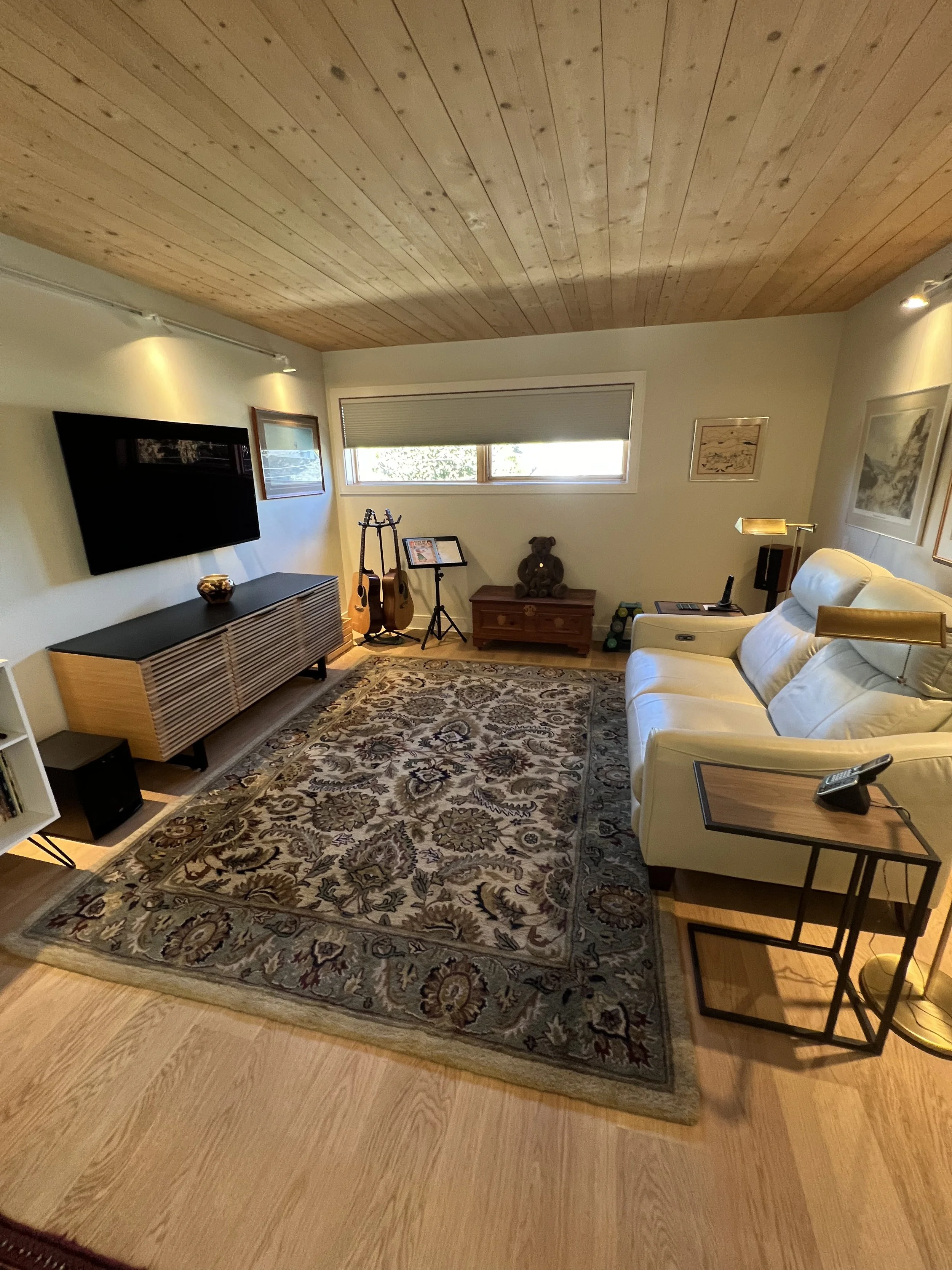 Living room with a patterned rug, white couch, wall-mounted TV, guitars, a small wooden table, framed artwork, a window with blinds, and a standing lamp.