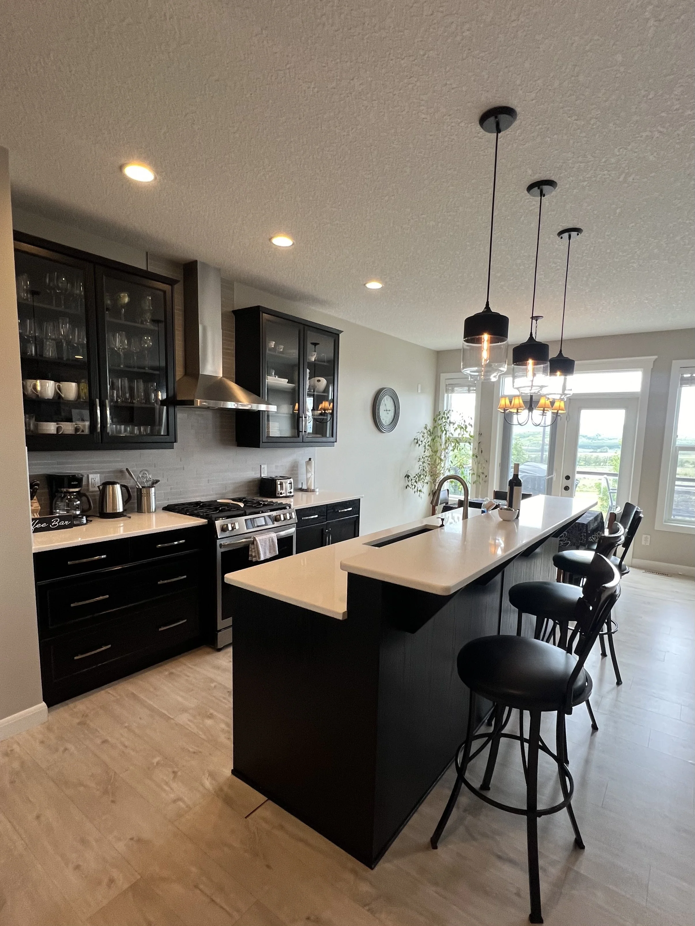 Modern kitchen with black cabinetry, pendant lights over a kitchen island, and a chalkboard-style sign reading 'Coffee Bar' on the countertop. There are bar stools around the island, and a large window and glass door leading outside. The kitchen is w