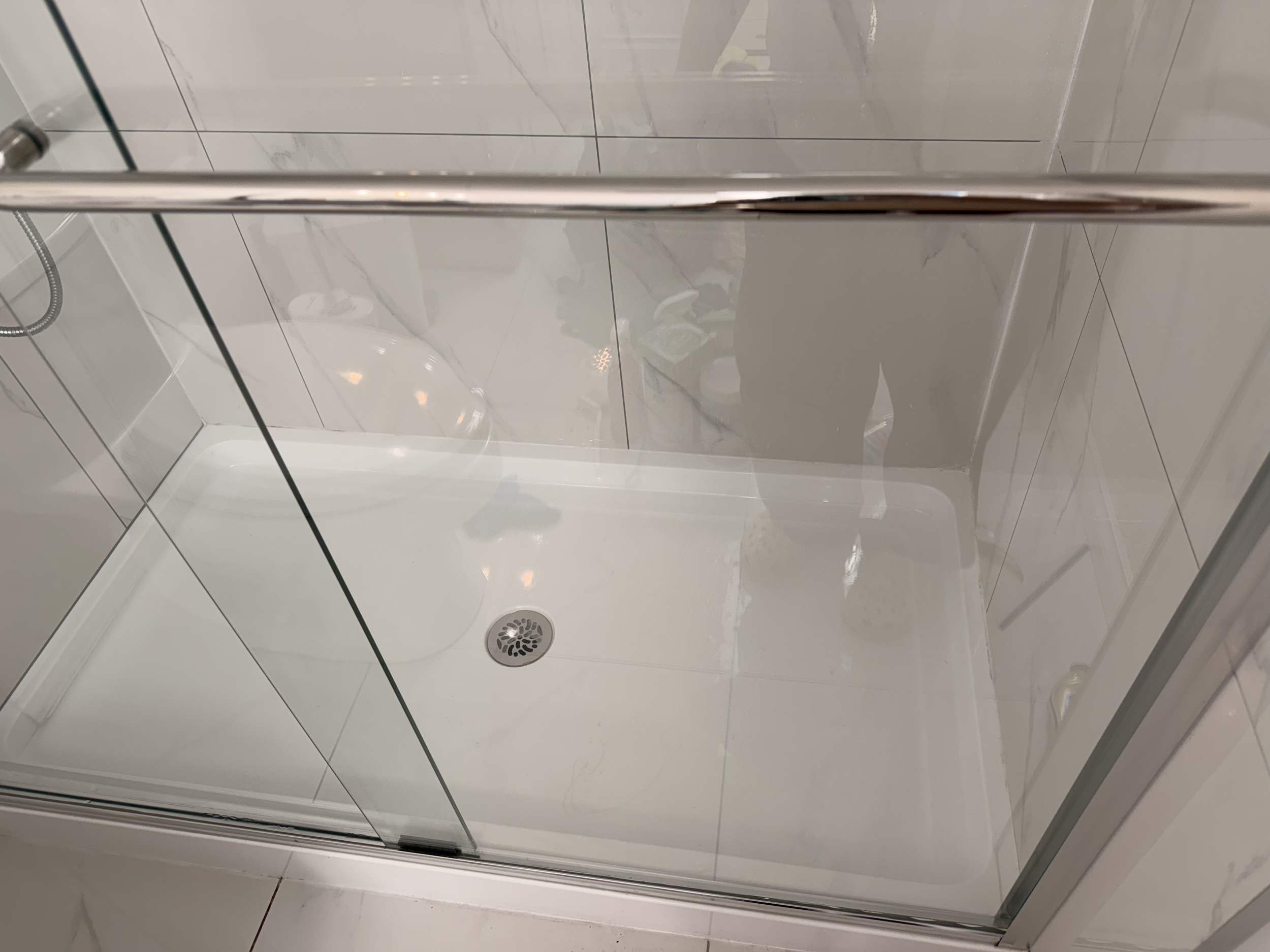 View of a shower area with white tiles, a glass door, a showerhead, and a drain on the floor.