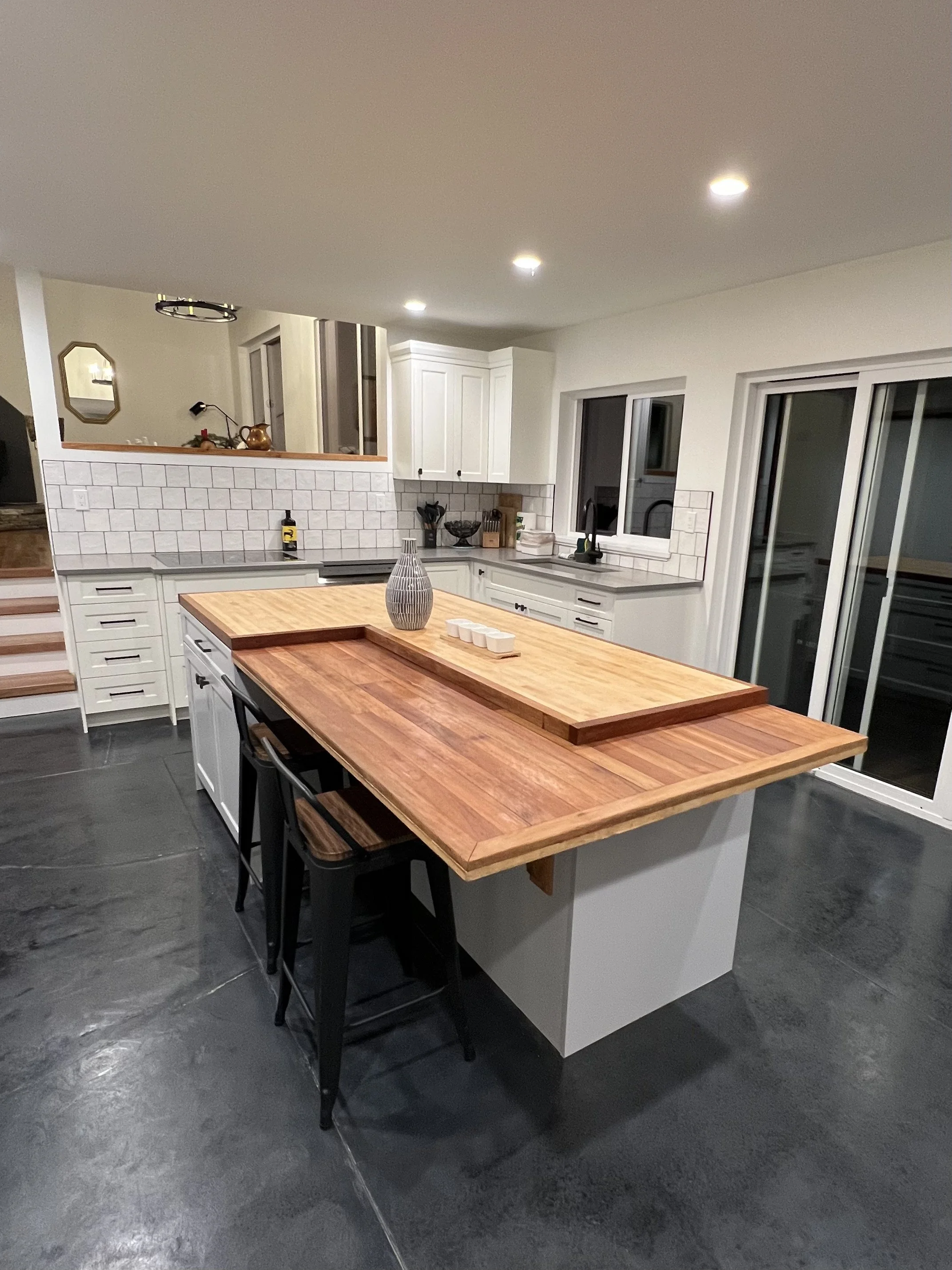 A modern kitchen with white cabinets, a wooden kitchen island, black chairs, gray countertops, and a white tiled backsplash. Large windows and sliding glass doors are visible, with recessed ceiling lights lighting the space.
