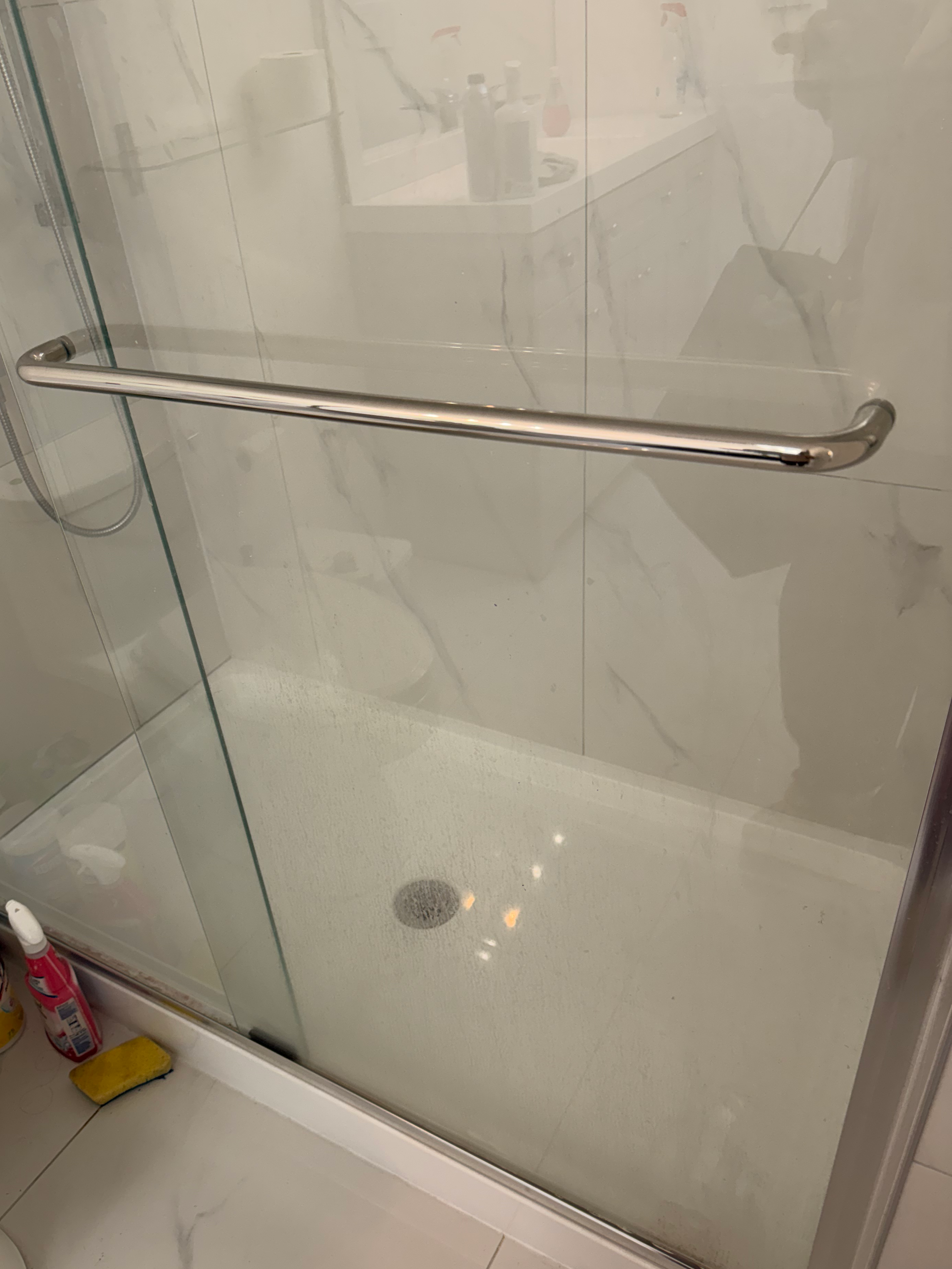 A glass shower door with a metal handle, reflecting a tiled bathroom floor, some bottles on a shelf, and bathroom cleaning supplies on the floor outside the shower.