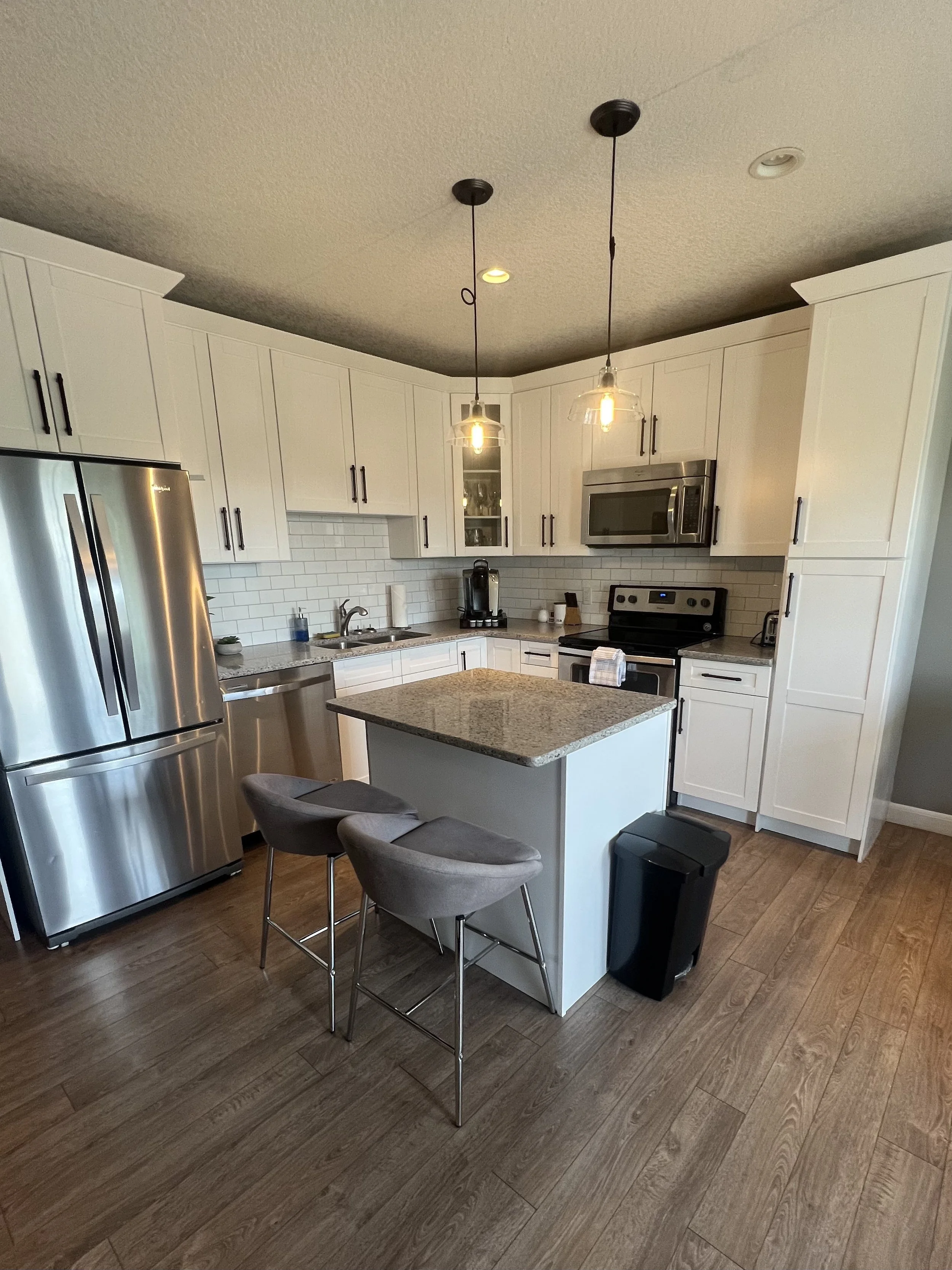 Modern kitchen with white cabinets, stainless steel appliances, granite island, two gray chairs, wood-look flooring, and pendant lighting.