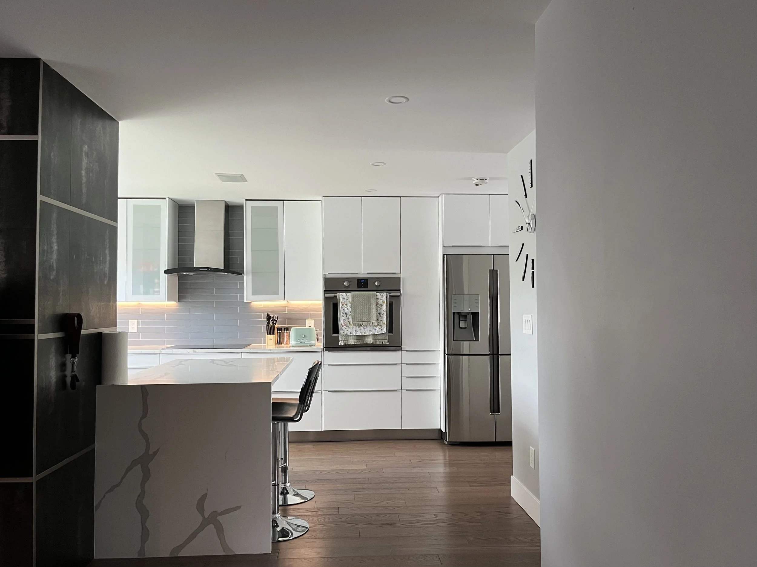 Modern minimalist kitchen with white cabinets, stainless steel appliances, gray backsplash, and hardwood flooring viewed from an open living space.