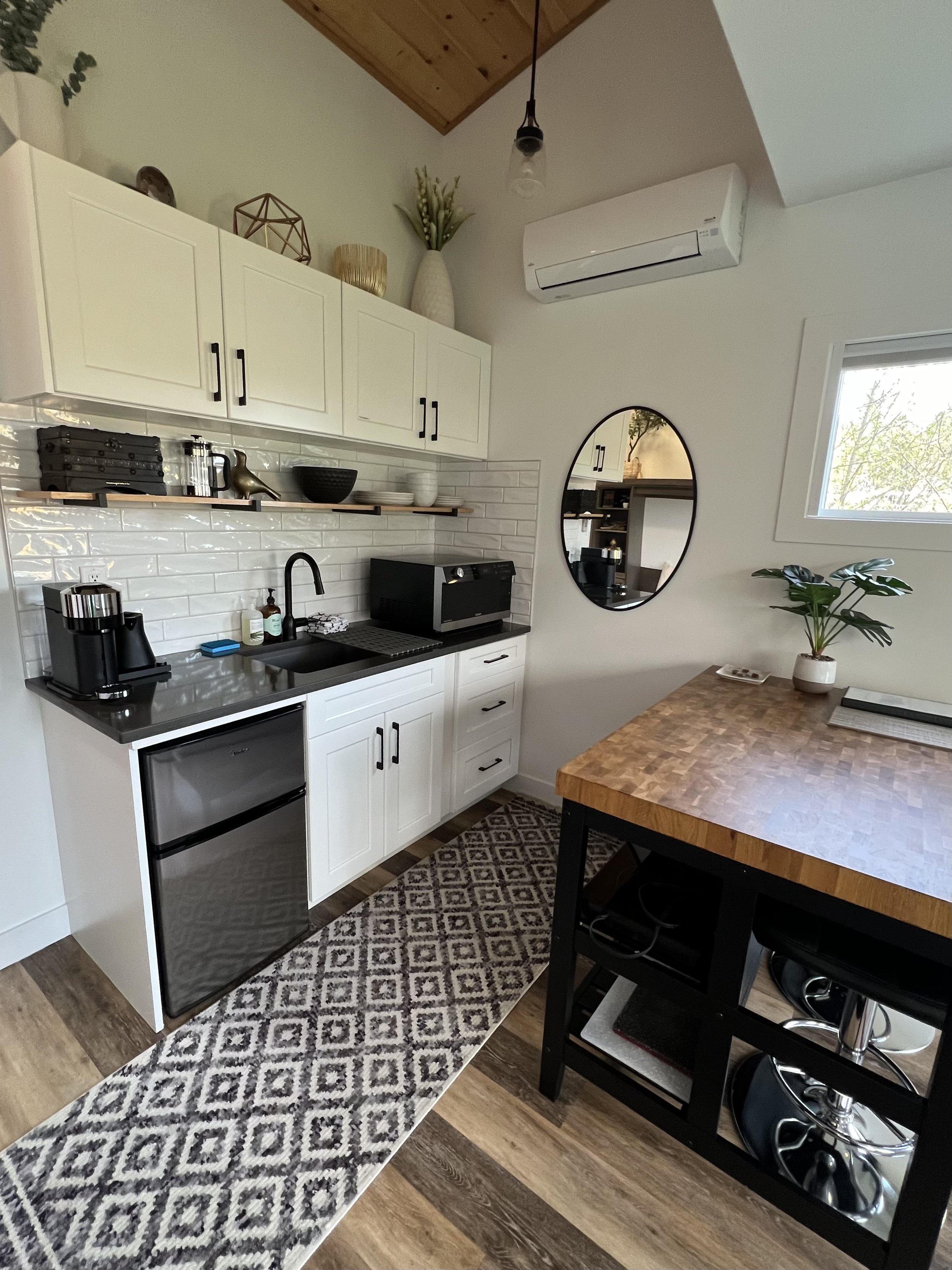 Kitchen area with white cabinets, black countertops, a small refrigerator, a microwave, and a coffee maker. There is a round mirror on the wall, a wooden table with a potted plant, a patterned rug, and a window with a view of trees outside. Decor inc