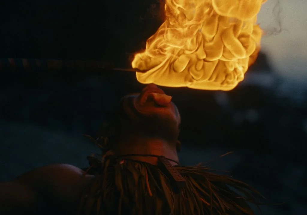 A person exhaling fire into the air during a fire dancing performance, with flames illuminating their face and surroundings.