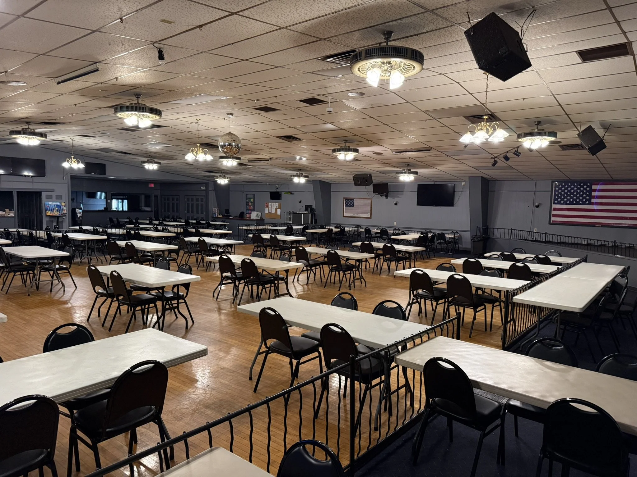 Large hall or banquet room with rows of empty rectangular tables and black chairs, decorated with a disco ball hanging from the ceiling, multiple ceiling lights, and a large American flag on the wall. There are several large flat-screen TVs mounted on the wall and some doors and notices in the background.