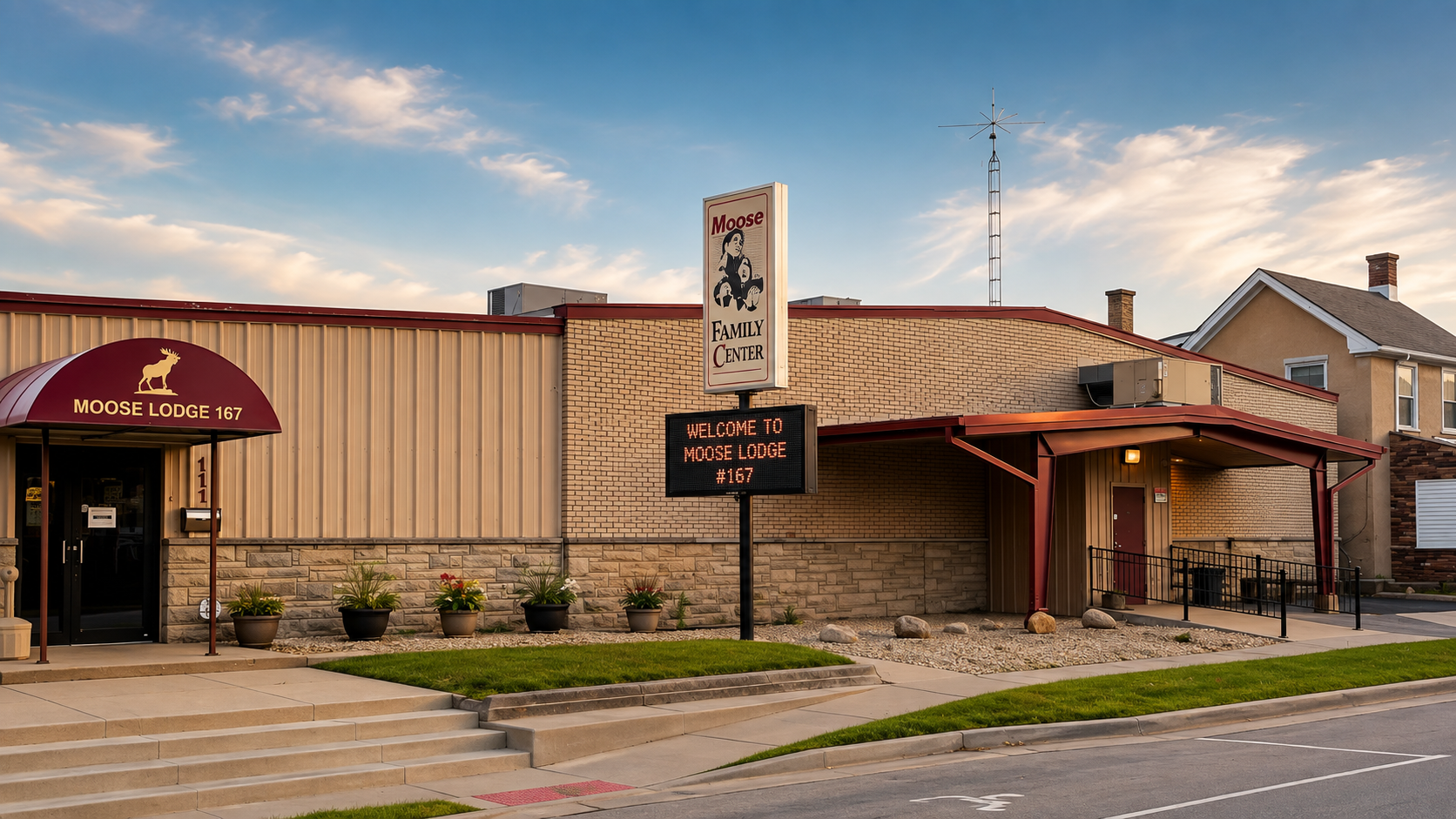 The exterior of Moose Lodge 167, a community facility with a brick and metal siding building, a red awning entrance, and a sign that reads 'Welcome to Moose Lodge 167'. There are potted plants near the entrance, a sidewalk, and a parking lot in front. The sky is partly cloudy.
